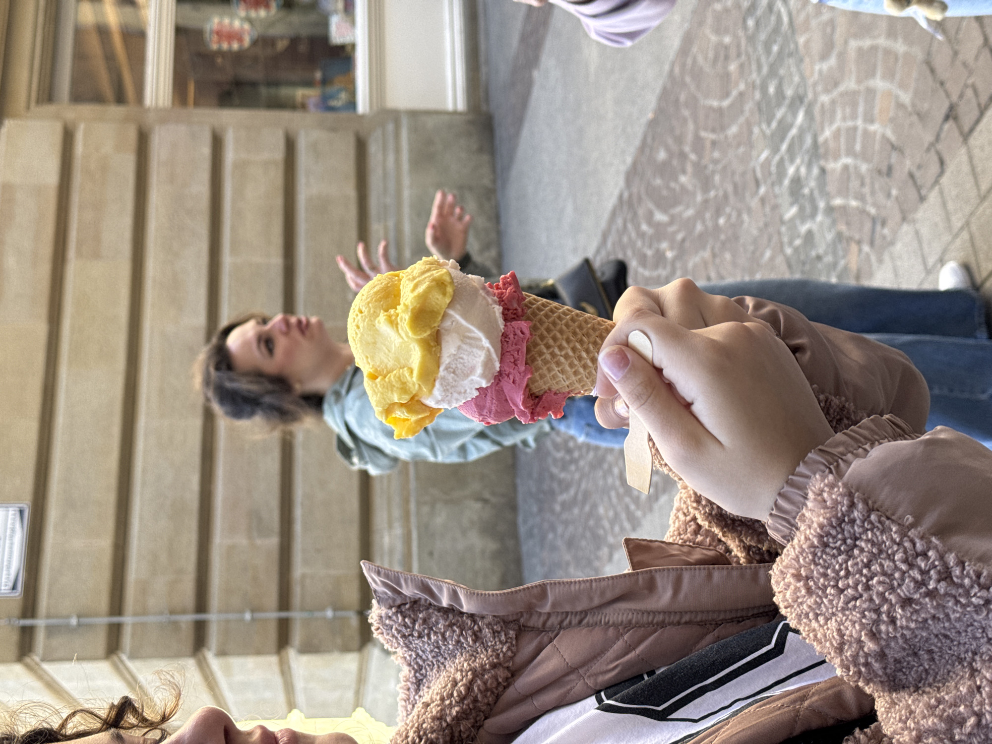 The image shows a person holding an ice cream cone with three scoops of ice cream: yellow, white, and pink. The person is wearing a brown jacket with a fleece collar. In the background, a woman in a green shirt stands near a beige building with vertical columns. The location is in the city of Luxembourg, Luxembourg, indicated by the architecture and street style. The scene appears to be taken outdoors, likely on a sidewalk.