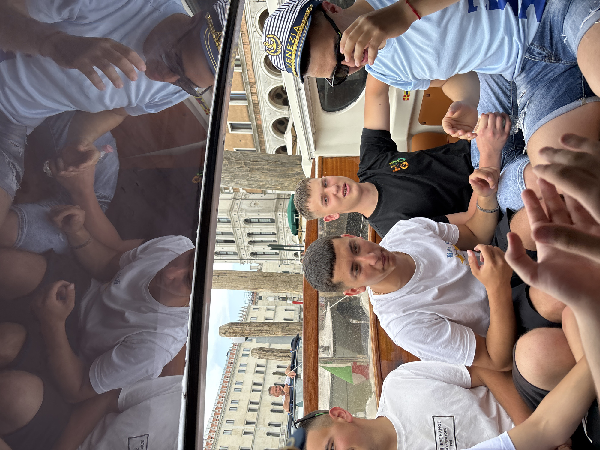 The image captures a group of young people on a boat in Venice, Italy. The boat appears to be passing along a canal, with the architecture of Venice visible in the background, including buildings with arched windows and ornate facades.
The group consists of at least five individuals. One person is wearing a striped "VENEZIA" cap and sunglasses. Several others are wearing light-colored t-shirts; one appears to be wearing a black t-shirt. The boat is moving through the water, and the reflections of the people and surrounding buildings are visible on a glass surface, likely a boat window or partition. One person can be seen steering another boat. The group seems to be looking at something in front of them, gesturing with their hands.