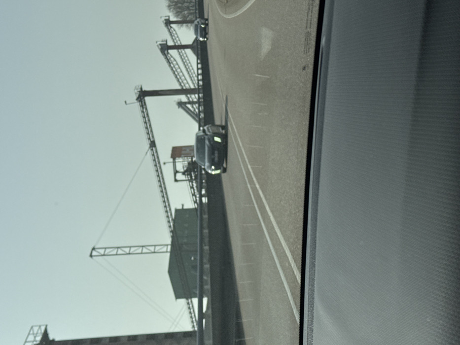 The image displays a forward view from within a vehicle, capturing an industrial landscape in Altenheim, Germany, specifically a gravel and sand processing plant likely located near Hauptstraße 55, 77743 Neuried-Altenheim. The vehicle's dark gray, textured dashboard occupies the lower portion of the frame. An asphalt road, delineated by white double solid lines and a partially visible white directional arrow in the right lane, extends into the mid-ground. The sky above is clear and light blue. Approaching on the opposing lane is a dark-colored hatchback, displaying illuminated daytime running lights. A second, less distinct dark-colored vehicle follows at a greater distance. The background is dominated by the industrial complex, characterized by multiple elevated metal conveyor belts supported by tall lattice structures. These belts connect various processing units, including a prominent green building or hopper and a tall, slender tower. A reddish-orange sign bearing the "UH" logo is affixed to one of the industrial structures. Bare-branched trees are visible on the far right horizon. No individuals are present in the scene.