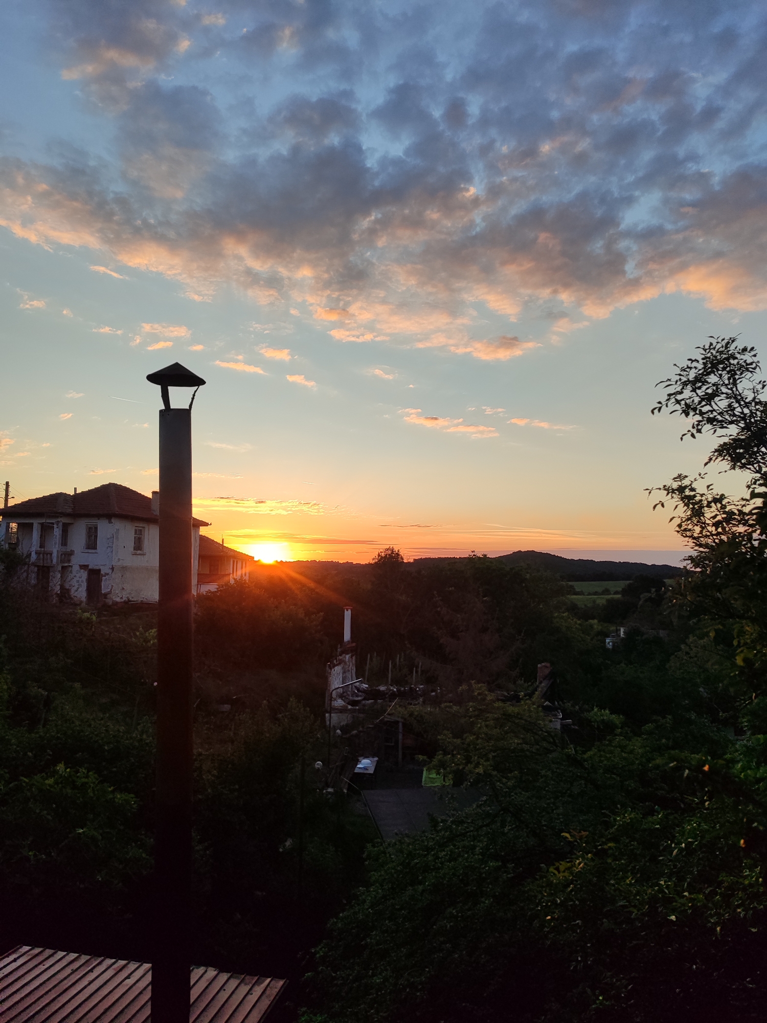 The image captures a sunset view from Varshilo, Bulgaria. In the foreground, a tall cylindrical metal chimney with a conical cap stands prominently. Below it, a glimpse of a corrugated metal roof can be seen. To the left, an aged two-story building with visible signs of disrepair, including crumbling plaster and multiple windows, is partially obscured by greenery.
The setting sun is positioned in the center of the image, radiating light rays. The sky is filled with clouds, with colors ranging from light blue to gray, tinged with orange and pink hues from the sunset.
The midground is filled with lush greenery, including trees and bushes that cover the surrounding terrain. Partially visible among the trees are houses or structures, indicating a settlement. The horizon is marked by hills in the distance.