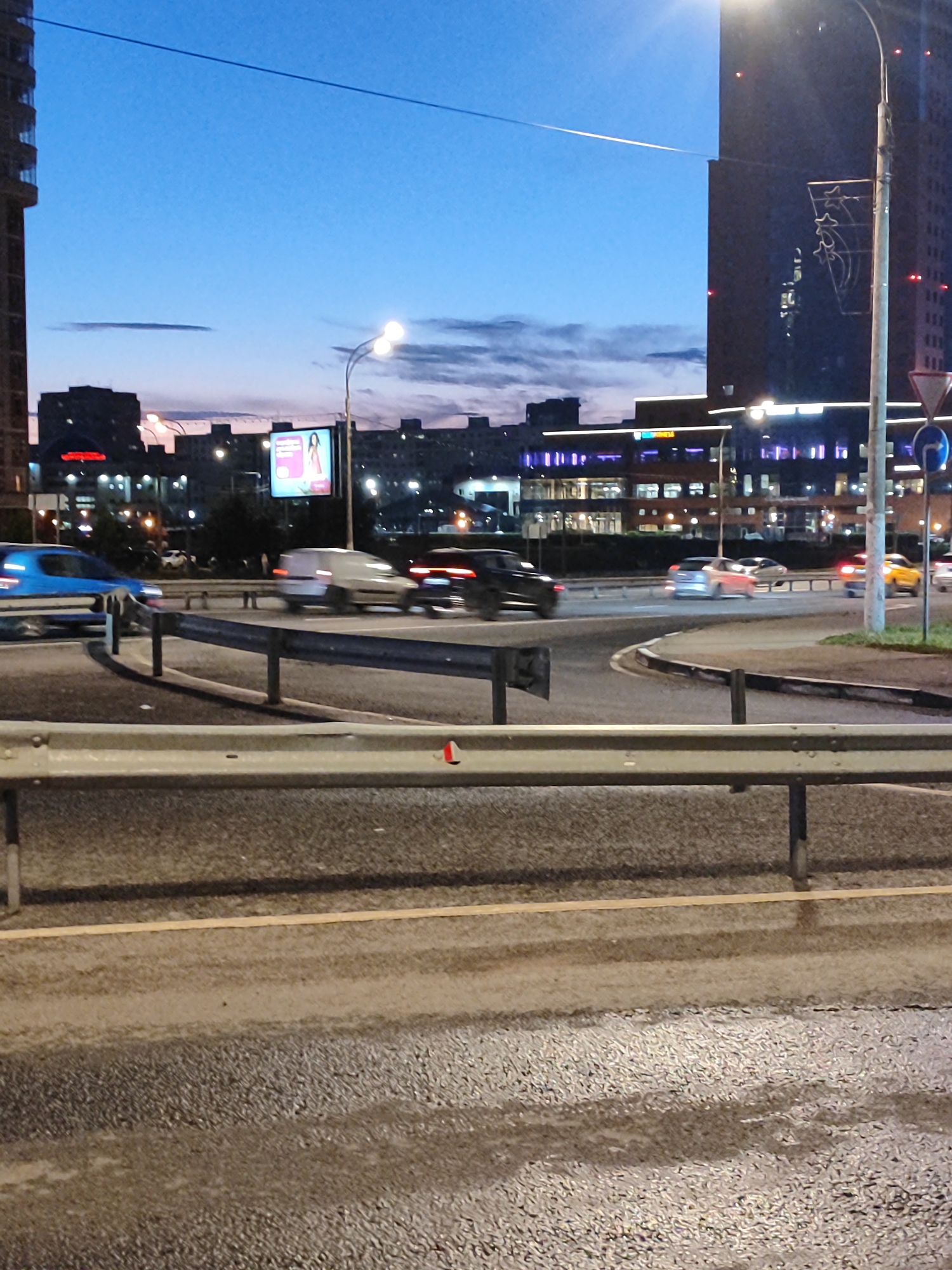 The image depicts a street scene at dusk in an urban environment. In the foreground, a wet asphalt road with visible lane markings is shown. A silver metal guardrail runs horizontally across the middle of the frame. In the background, a highway intersection is visible with several cars in motion, their headlights and taillights blurred due to the long exposure or motion. Buildings, illuminated with lights, line the street in the distance. The sky is a deep blue, transitioning to lighter shades near the horizon, indicating either sunrise or sunset. There is a billboard with an advertisement on it. The scene appears to be a typical city view during twilight hours.