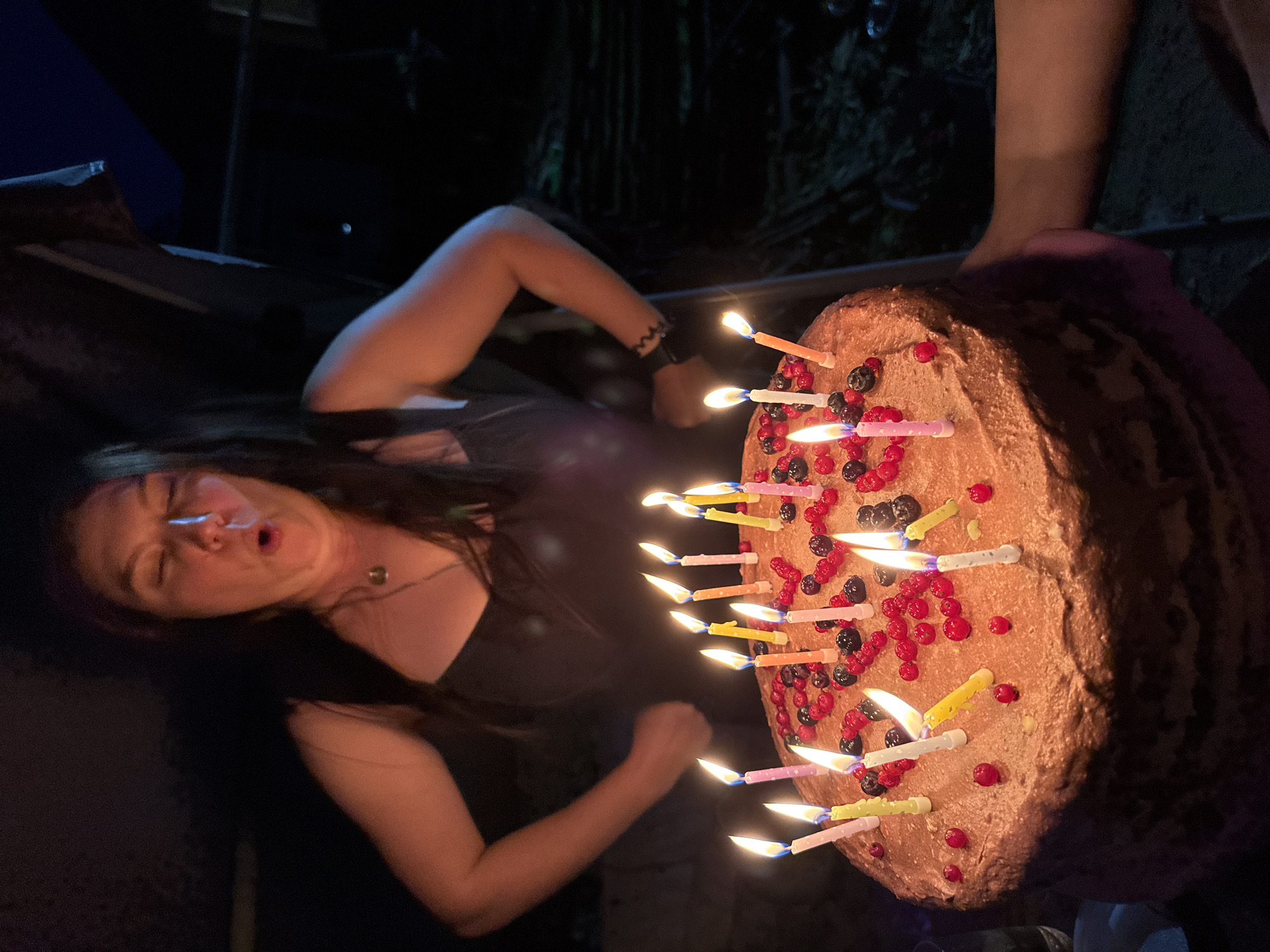 A woman with her eyes closed and mouth open is blowing out candles on a chocolate cake decorated with berries and dots. She has dark hair and is wearing a black dress and a pendant necklace. The cake is lit by numerous colorful candles. The scene appears to be outdoors at night, with dark foliage in the background. There are hands visible on the right side of the image, suggesting other people are present. The location is specified as Хохряки, Russia.