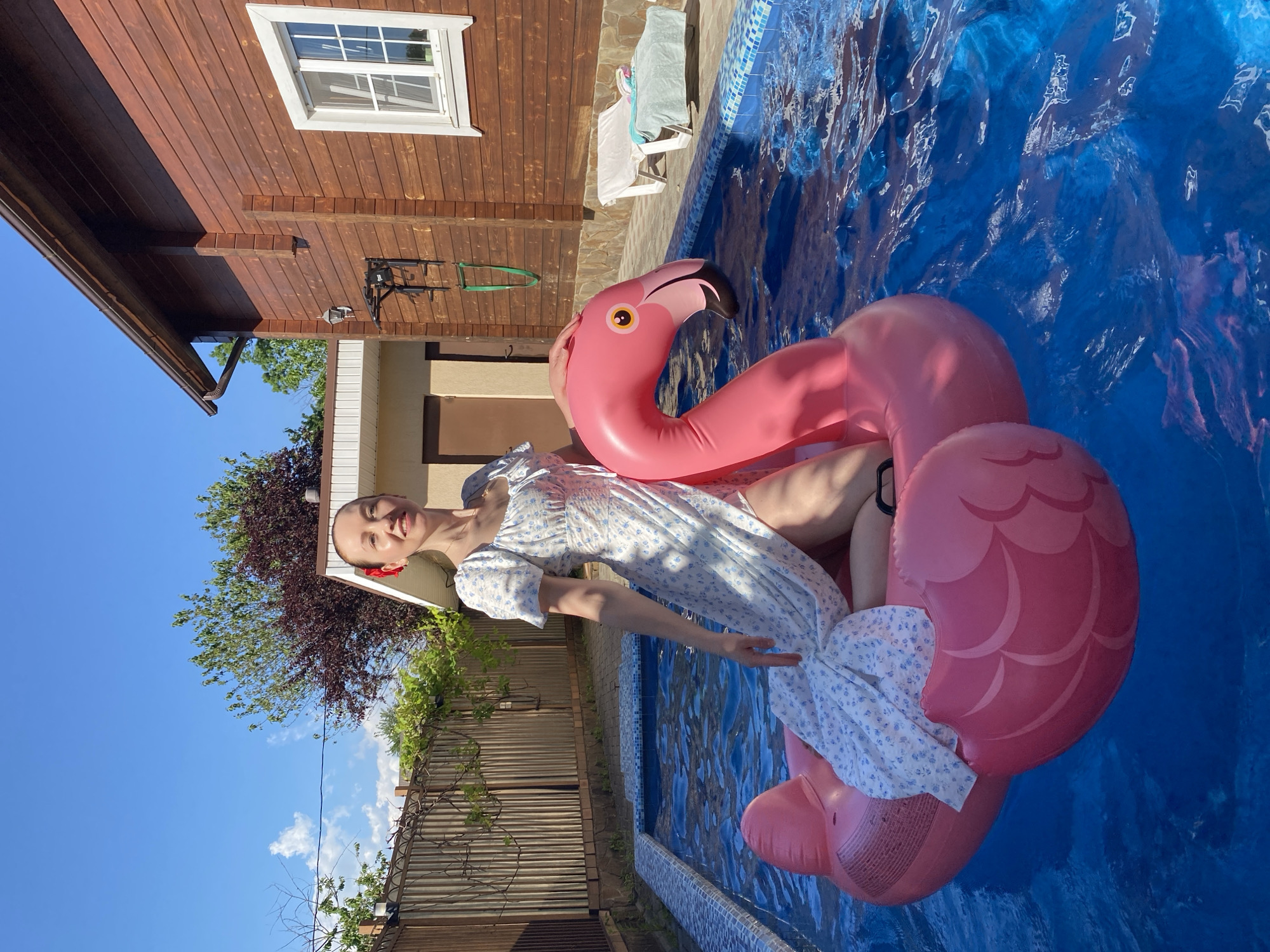The scene depicts a woman in an outdoor swimming pool, seated on a large pink inflatable flamingo float under bright daylight in the city of Dagomys, Russia. The woman, appearing to be in her 20s or 30s, has fair skin and light hair, accessorized with a small red ornament on the right side of her head. She wears a white midi-length dress with a light blue floral pattern, featuring puff sleeves, a square neckline, and a high slit revealing her right leg. She is smiling faintly and looking directly towards the viewer, with her left hand resting on the flamingo's head.
The inflatable flamingo is predominantly pink, with a black beak, a yellow eye with a black pupil, and white highlights. The pool water is bright blue, exhibiting ripples and reflections, and its edge is lined with blue and white mosaic tiles.
The background features various structures and vegetation. To the left, a wooden fence with a dark ornate metal top section is covered with green vines. Beyond the fence, a clear blue sky with sparse white clouds is visible, along with green and reddish-brown foliage from trees. Adjacent to the fence and behind the pool, a light-colored building with a brown door and a dark brown roof trim is present. To the right of this building is a larger house with dark brown horizontal wooden siding. This house features a dark brown gutter system, a wall-mounted light fixture, and an outdoor exercise pull-up bar attached to its wall. A white-framed window with multiple panes is visible on the right side of the wooden house. A paved area surrounds the pool, and two white lounge chairs are positioned to the right of the pool, one draped with a light blue towel. The strong sunlight casts clear shadows.