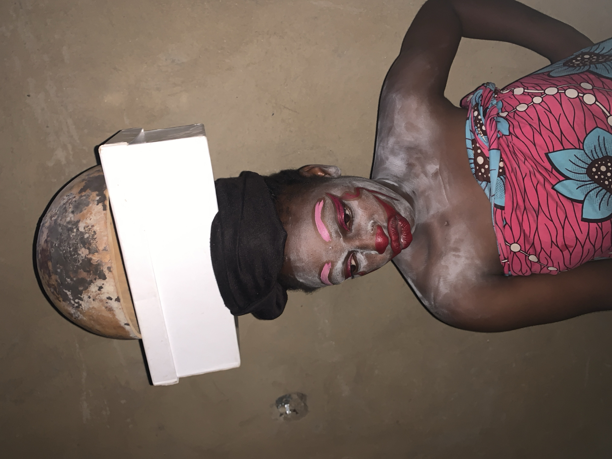 In Agban, Nigeria, a person with dark skin is posed against a plain, earthen-colored wall. They are wearing an elaborate face paint design, with white powder covering their face and chest, bright pink eye shadow, and dark red lipstick applied in a clown-like fashion. A black headwrap is worn, and over this, a white box is balanced, topped by a round, textured object resembling a gourd or a weathered bowl. The individual is draped in a brightly colored fabric featuring a pink background with blue floral patterns and black lines. The lighting is dim, creating shadows and highlighting the facial makeup. The setting appears to be indoors, possibly a dwelling, given the wall texture and the indoor nature of the objects.