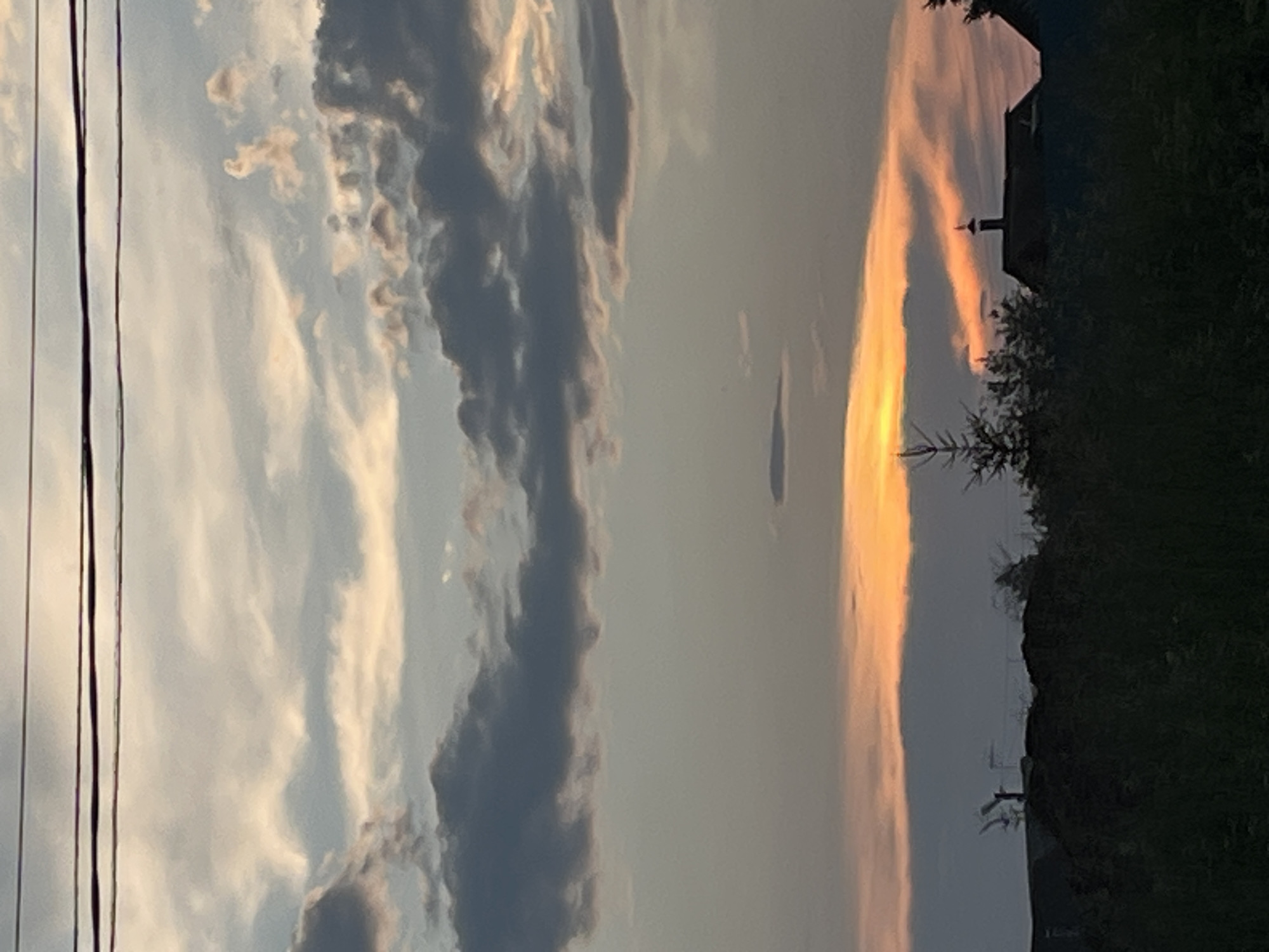 This is a vertical, medium-range photograph of a sunset over a rural area, likely in Ludorvay, Russia. The sky dominates the upper two-thirds of the frame, filled with a mix of grey, white, and orange clouds. Three thin, dark power lines stretch horizontally across the top of the image. The lower third of the image shows a silhouetted landscape of trees and rooftops against the brightly lit sky. One prominent building has a chimney with a small, pointed cap. A thin, spiky evergreen tree stands to the left of the building. The overall impression is one of a peaceful, twilight scene.