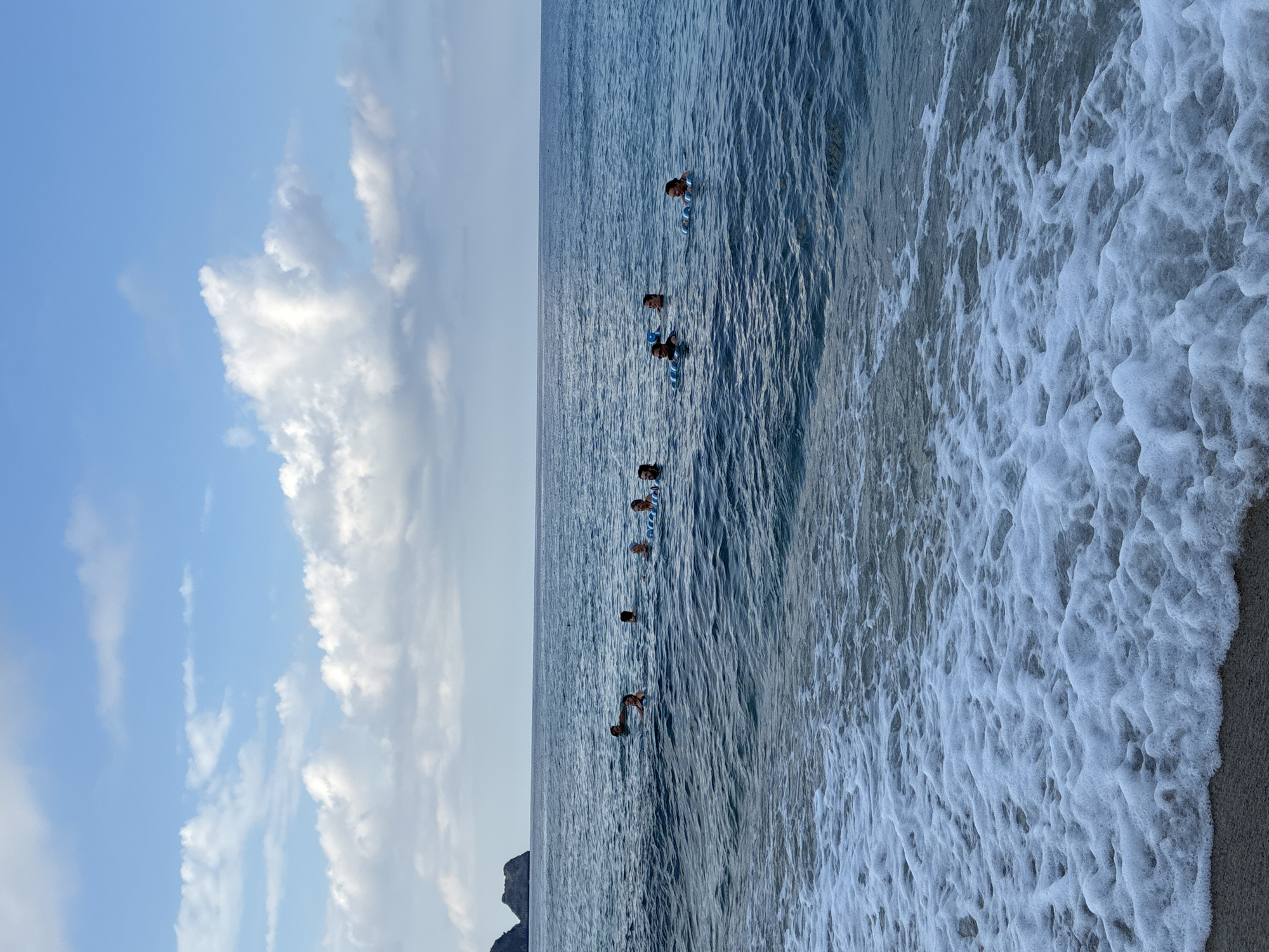 A group of approximately eight people are swimming in the sea with flotation devices. The water is a deep blue with moderate waves. The sky is blue with scattered white clouds. In the distance, a rocky island is visible on the left. The scene appears to be a beach or coastal area. The overall setting is a natural environment, likely the Mediterranean Sea. The presence of flotation devices suggests a recreational activity or training.