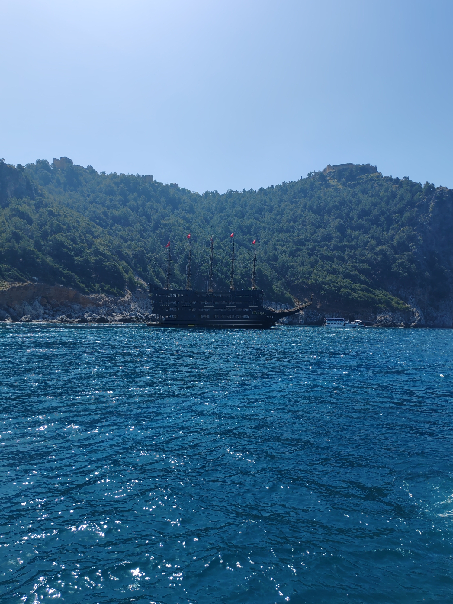 The image shows a view of the sea near Alanya, Turkey. A large, dark-colored pirate ship with red flags is anchored near a coastline covered with green vegetation and rocky outcrops. In the distance, on top of the hill, are what appear to be ruins or castle fortifications. The sea is a deep blue color with sunlight reflecting off the water. Another white boat can be seen near the pirate ship, closer to the coastline. The sky is clear and blue.