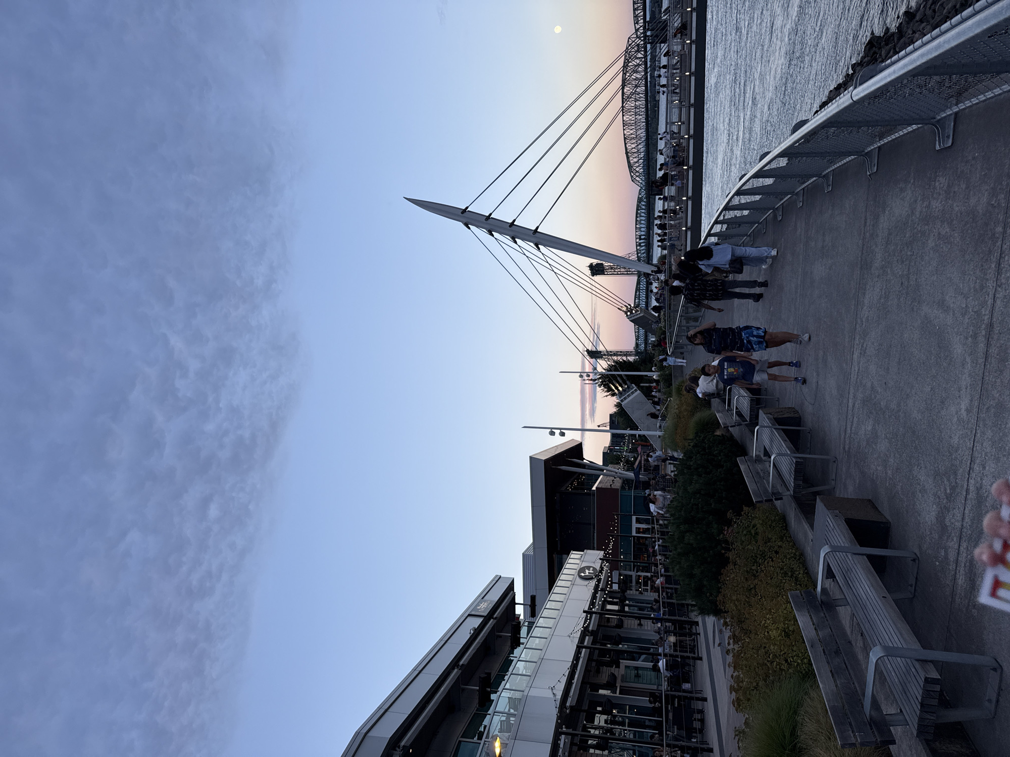 The image depicts a waterfront promenade in Vancouver, Washington, USA, during what appears to be early evening. The sky is a gradient of blue and pink, with scattered clouds. On the left side of the frame, a modern building with large windows and outdoor seating areas is visible, likely housing restaurants or shops. String lights adorn the building's exterior.
A paved walkway runs along the water's edge, populated by pedestrians. Several individuals are walking, including a young boy and a young girl walking together, and a group of adults. To the right of the walkway, the Columbia River flows, with rocky embankments along the shore. In the distance, a striking modern pedestrian bridge with a distinctive angled pylon and cable-stayed design spans the river. Beyond this bridge, a more traditional truss bridge is also visible. The overall scene suggests a lively public space at dusk, with people enjoying the waterfront.