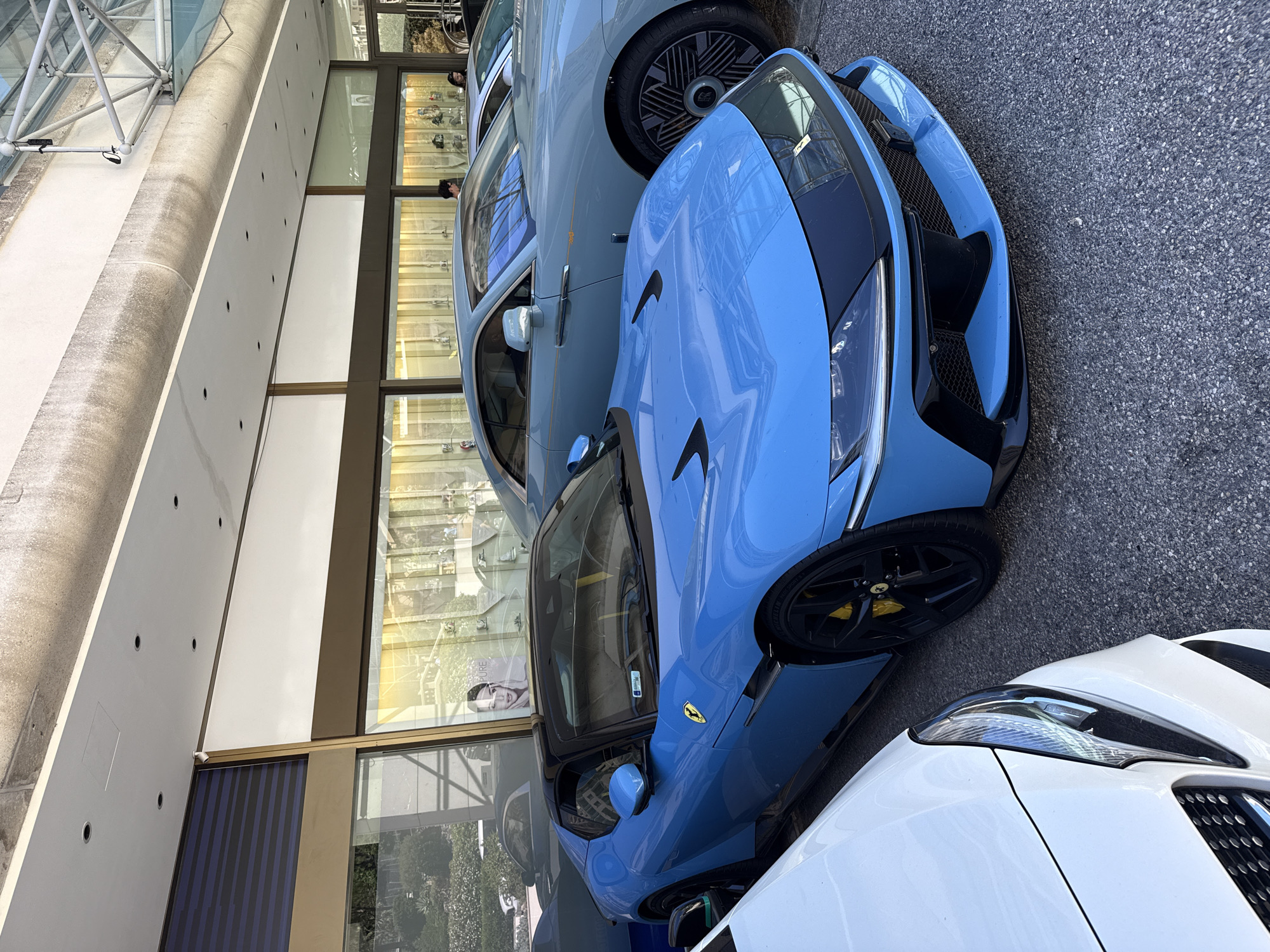 In Monaco, Monaco, a light blue Ferrari Roma is parked outside a building with a beige facade and large windows. The Ferrari is the most prominent subject, with its black convertible top, black accents, and yellow brake calipers visible. To the right of the Ferrari is a light blue Rolls-Royce convertible, also with a black top. Partially visible on the left is the white front of another vehicle. The building's facade features rows of lights and display cases, suggesting it might be a luxury retail establishment. The ground is paved with asphalt. Two individuals are visible through the building's windows. The lighting suggests it is daytime.
