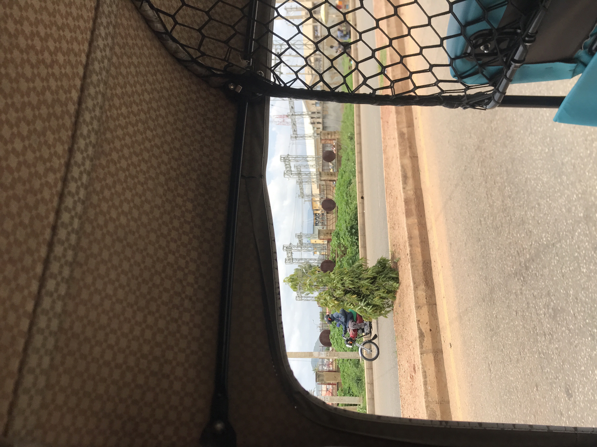 The image depicts an outdoor urban or semi-urban scene, viewed from the interior of a vehicle, likely a tricycle, looking outwards over a roadway. The setting is in the city of New Stadium, Nigeria.
In the immediate foreground, the overhead interior roof of the vehicle is visible, covered in a light brown fabric featuring a repeating, small-scale checkerboard or houndstooth pattern in a darker brown. To the right, a black hexagonal mesh screen or net extends downwards, partially obscuring the view. Below this mesh, a segment of bright turquoise fabric, possibly a seat cushion, is visible.
The midground features a light grey asphalt road, seemingly two lanes wide, traversed by a red motorcycle moving from left to right. The motorcyclist wears a dark helmet and a blue jacket, with a light-colored item visible on their back. A raised, light brown concrete curb separates the road from a narrow, unpaved median strip. Within this median, a small, conical green tree with dense foliage is situated.
In the background, a series of prominent grey, industrial-style lattice structures, characteristic of an electrical substation or high-voltage power lines, are dominant. Behind these structures, light-colored buildings are discernible. To the far left, faint hills or mountains are visible on the horizon. Several large, dark, circular objects, resembling industrial containers, are present on the ground behind a low barrier in the left background. Further right, past the interior mesh, blurred figures of people and a small white canopy or umbrella are visible in the distance. The sky is overcast with varying shades of grey clouds, indicating a cloudy day with diffused lighting.