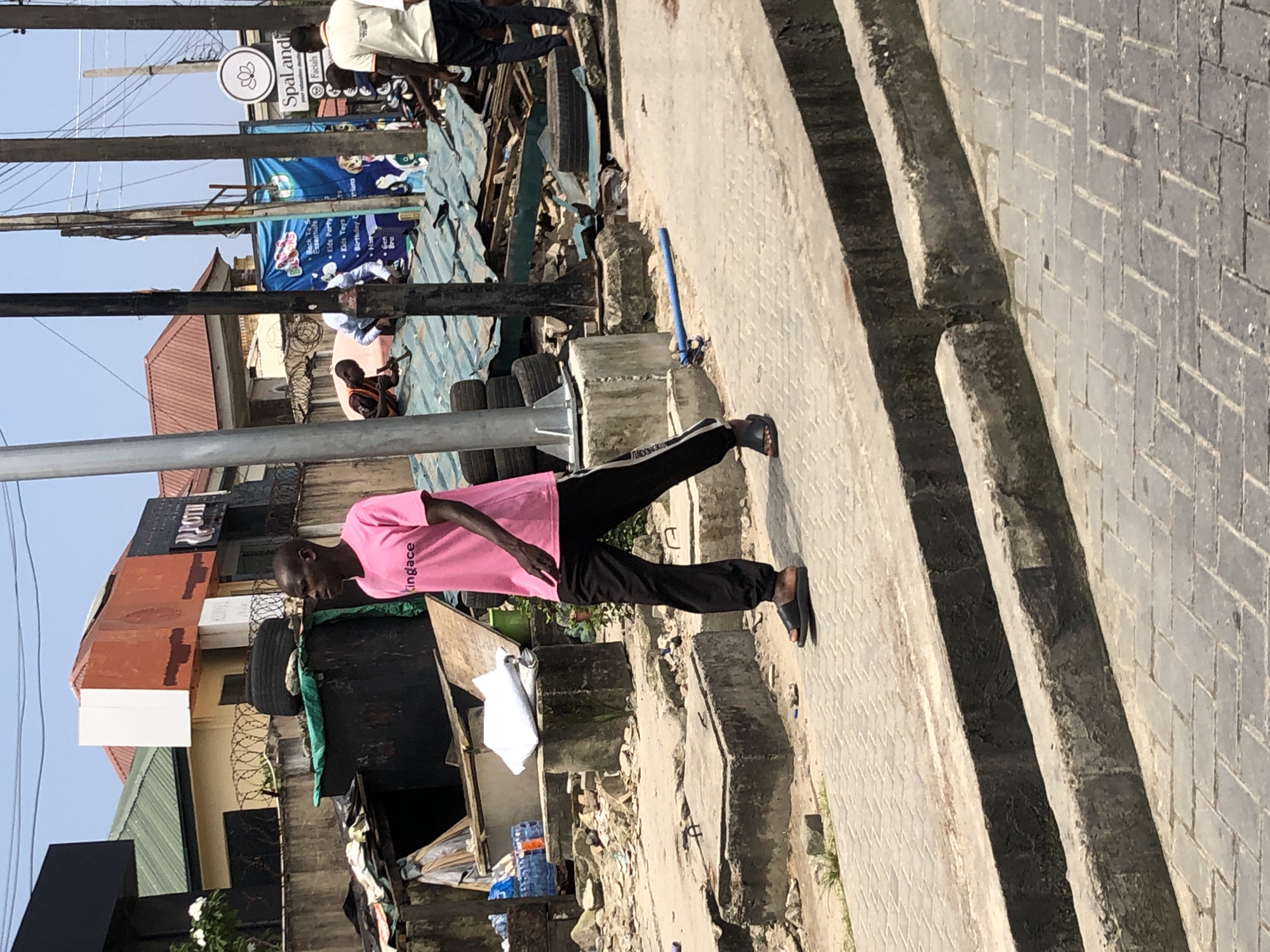 The image depicts an outdoor scene in Moba, Nigeria, likely during the daytime. The primary subject is a Black man wearing a bright pink t-shirt with the word "kingace" printed vertically on the back, and black pants with a white stripe down the leg. He is wearing black flip-flops. He is walking on a paved sidewalk next to a concrete structure. The background shows a cluttered street scene with various objects including tires, construction debris, corrugated metal roofing, and what appears to be a building with red and brown siding. A blue sign with a lotus flower logo and the text "Spaland" is visible in the upper left corner, along with another blue banner featuring cartoon characters. The overall atmosphere suggests a bustling, possibly commercial or residential area, with signs of ongoing construction or repair. The lighting indicates it is daytime.
