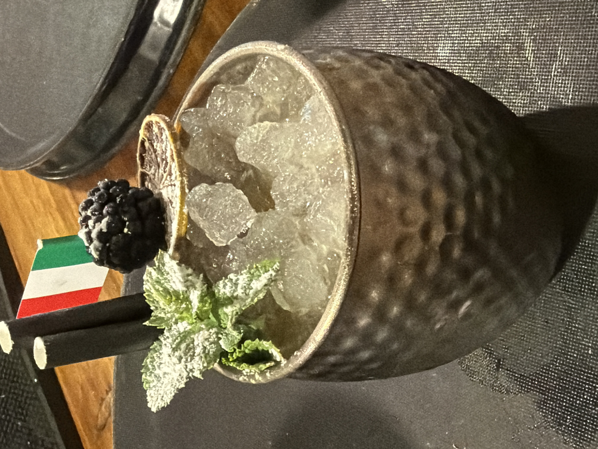 A close-up, top-down shot features a dark, textured ceramic glass filled with crushed ice. A blackberry rests on the rim, next to a dehydrated orange slice. Fresh mint sprigs, dusted with powdered sugar, are nestled amongst the ice. Two black straws are placed in front of the glass, and a small Italian flag is tucked behind the blackberry. The glass sits on a dark, textured surface, possibly a placemat or cloth, with a wooden surface visible to the left. The lighting suggests an indoor setting, likely a bar or restaurant. No people are visible. The scene is static, with no discernible activity beyond the preparation or presentation of the drink. The overall impression is one of a crafted beverage, ready to be served.