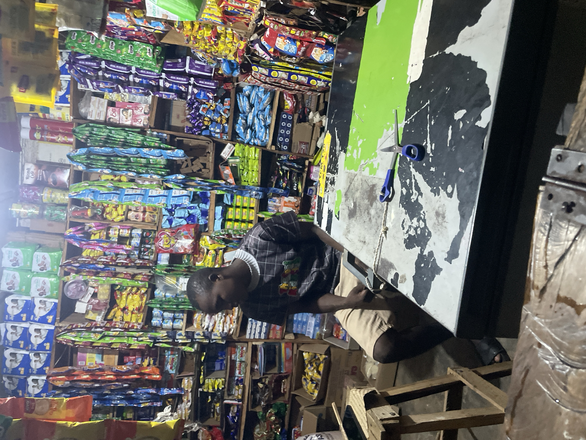 A young Black man is seated in a Nigerian convenience store, likely Lokoja. He wears a black and gray checkered t-shirt and light brown shorts, with black sandals on his feet. The store is packed with shelves full of merchandise including baby diapers, snacks, drinks, and toiletries. In the foreground, a table is covered with a checkered black and green cloth, with a pair of blue-handled scissors resting on it. The man is looking to his left, seemingly engaged in thought or observing something outside the frame. The lighting is dim, creating shadows throughout the store.