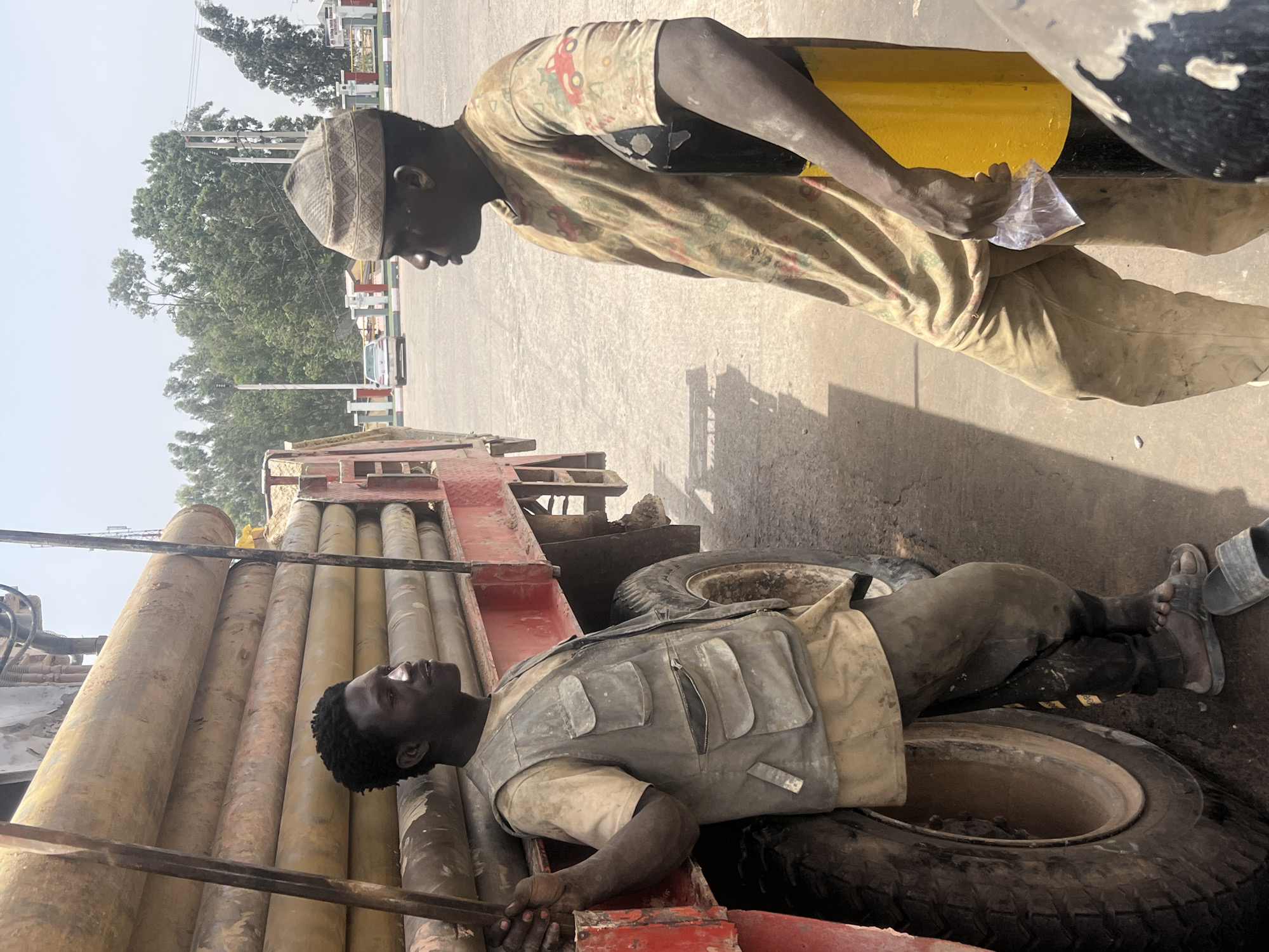 The scene depicts two men in an industrial setting, potentially in Jos, Nigeria. One man lies horizontally on a red flatbed truck, partially resting on a tire and amongst stacked pipes. He wears a light vest over a light-colored shirt. The second man stands on the ground in a patterned shirt, wearing a hat. He appears to be holding a small plastic bag. The location appears to be outdoors with buildings and trees in the background.