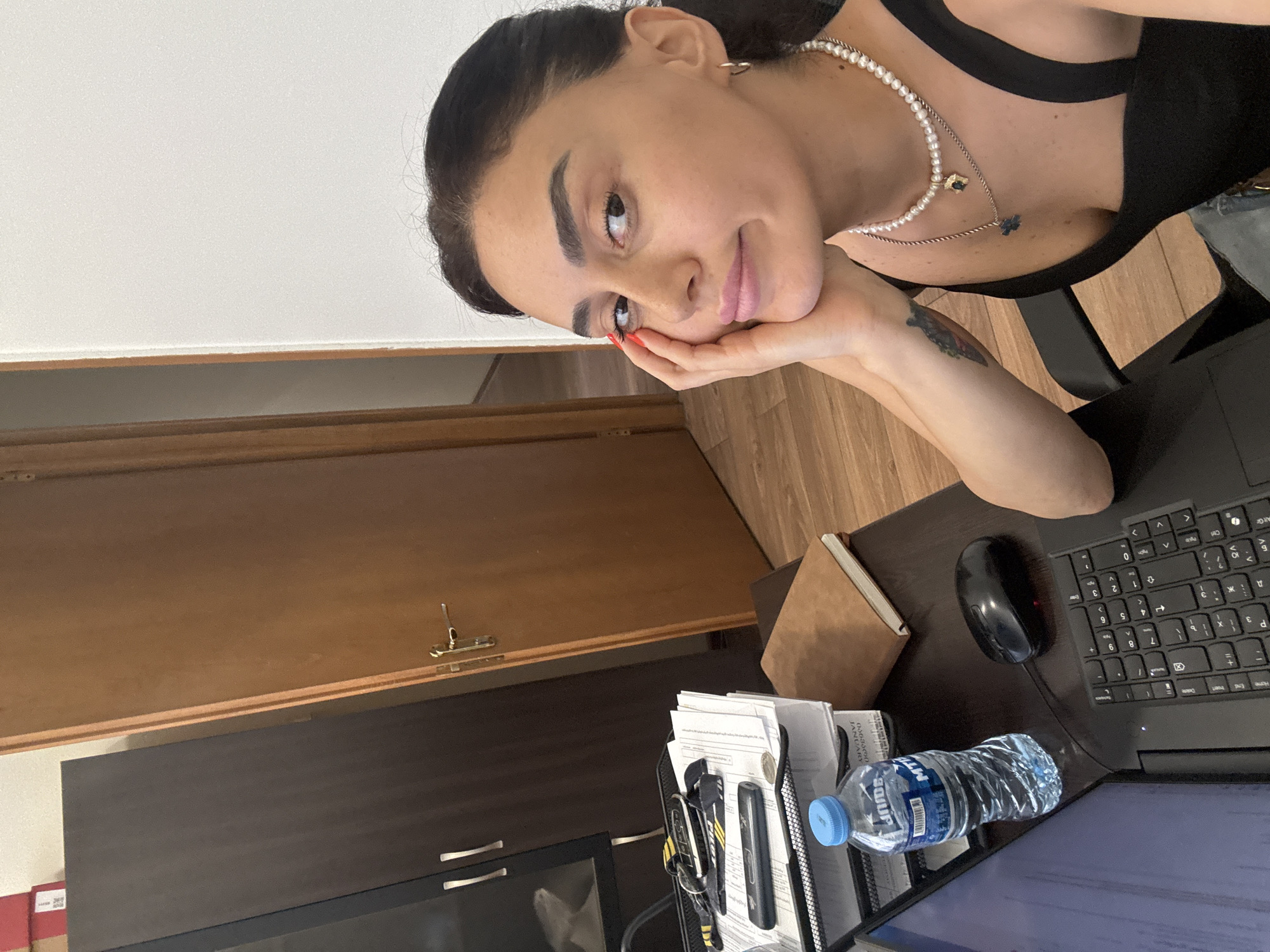 A medium shot shows a young woman with dark hair and brown eyes, resting her chin on her hand. She is wearing a black tank top and a pearl necklace. She is seated at a dark wooden desk, with a laptop, a bottle of water, a computer mouse, and a stack of papers in front of her. To her left is a dark brown cabinet with silver handles. Behind her, a light brown wooden door stands ajar. The floor is a light wood laminate. The overall impression is of a home office environment. The image does not contain enough information to determine the specific location within Tbilisi, Georgia.