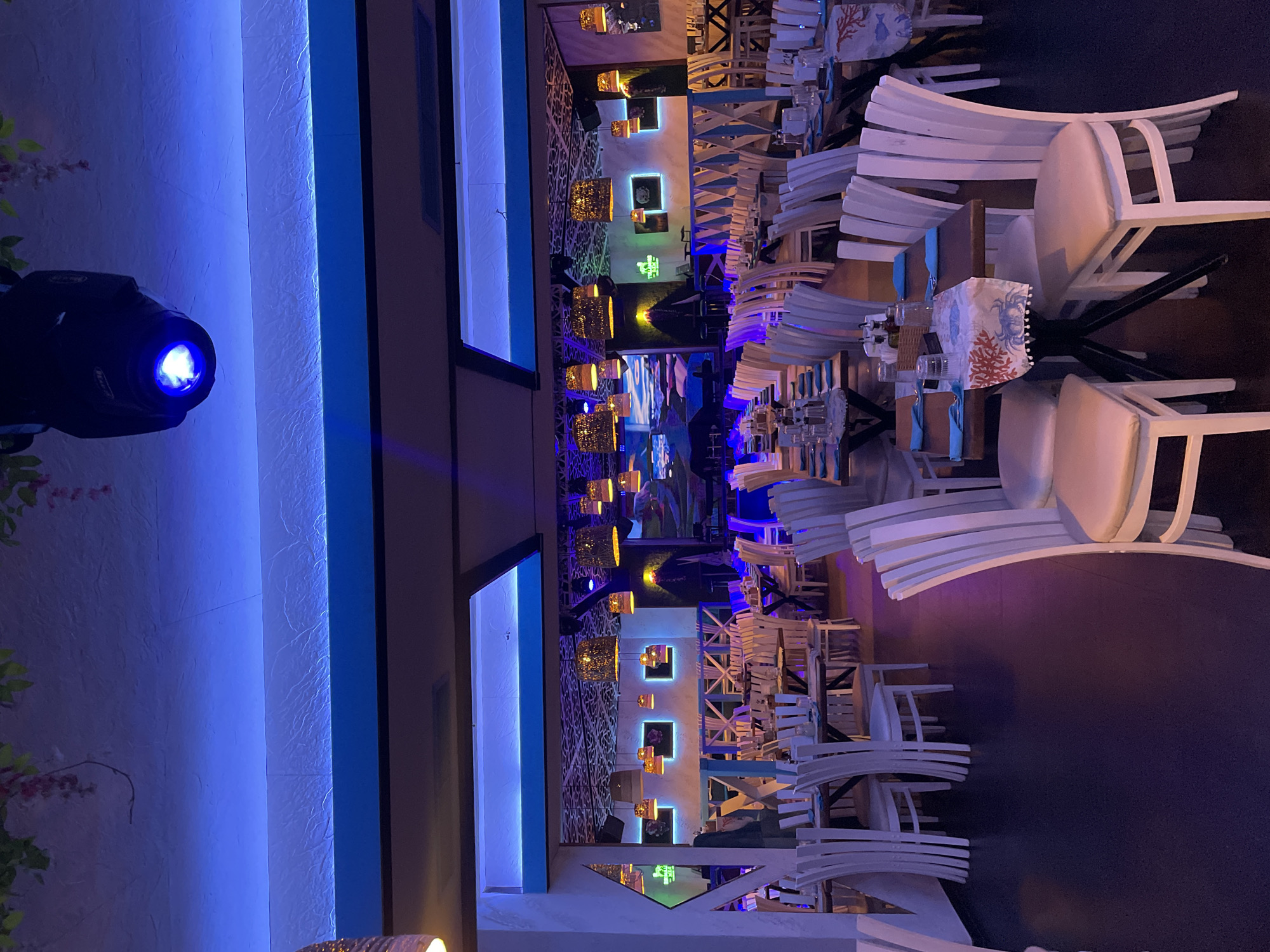The image shows the interior of a restaurant or bar in Plovdiv, Bulgaria. The space features modern design elements with a predominant blue and white color scheme. Tables are set with chairs stacked atop them, likely at the end of service, with white frames and upholstered seats. Decorative lights hang from the ceiling, adding warmth to the cool-toned atmosphere. A lighting fixture in the foreground casts a bright blue glow, further enhancing the ambiance. Artwork and mirrors adorn the walls, contributing to the overall aesthetic. No people are visible in the shot.