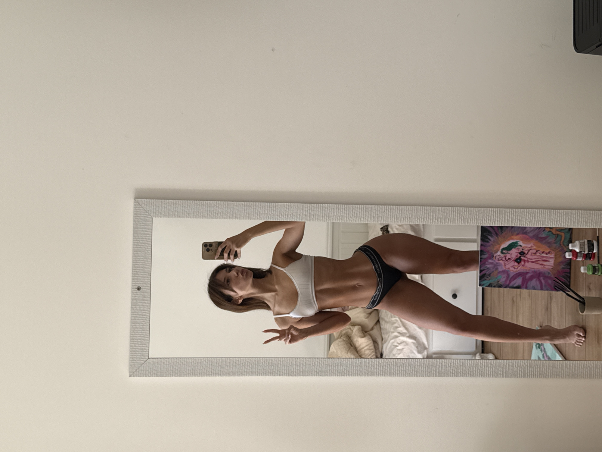 A woman in a white sports bra and black underwear takes a selfie in a mirror. She is holding a smartphone in her right hand and making a peace sign with her left. The background visible in the mirror shows a bed, a white dresser, a painting, and some art supplies on a wooden floor. The mirror is hanging on a white wall. No information is available to suggest a location in Sofia, Bulgaria.