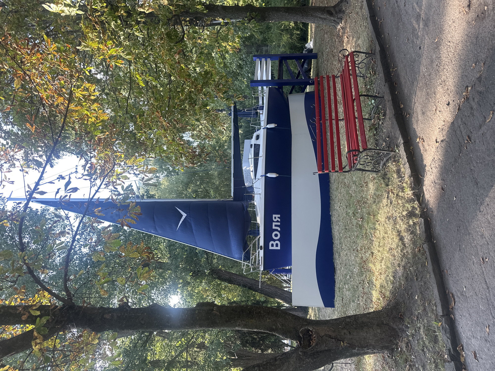 The image depicts a stationary, mid-sized sailboat that has been placed on land in a park-like setting. The boat is painted white and blue, with the word "Воля" (Volya) prominently displayed in white lettering on the blue hull. A large, dark blue sail is raised, featuring a white bird-like emblem. The sailboat is situated on a grassy area, surrounded by mature trees with dappled sunlight filtering through their leaves. In the foreground, a bright red park bench with ornate black metal legs sits on the edge of an asphalt pathway. The overall scene suggests a decorative or artistic installation rather than a functional vessel. This installation is located in Ternopil, Ukraine, specifically in the Ternopil Pond area.