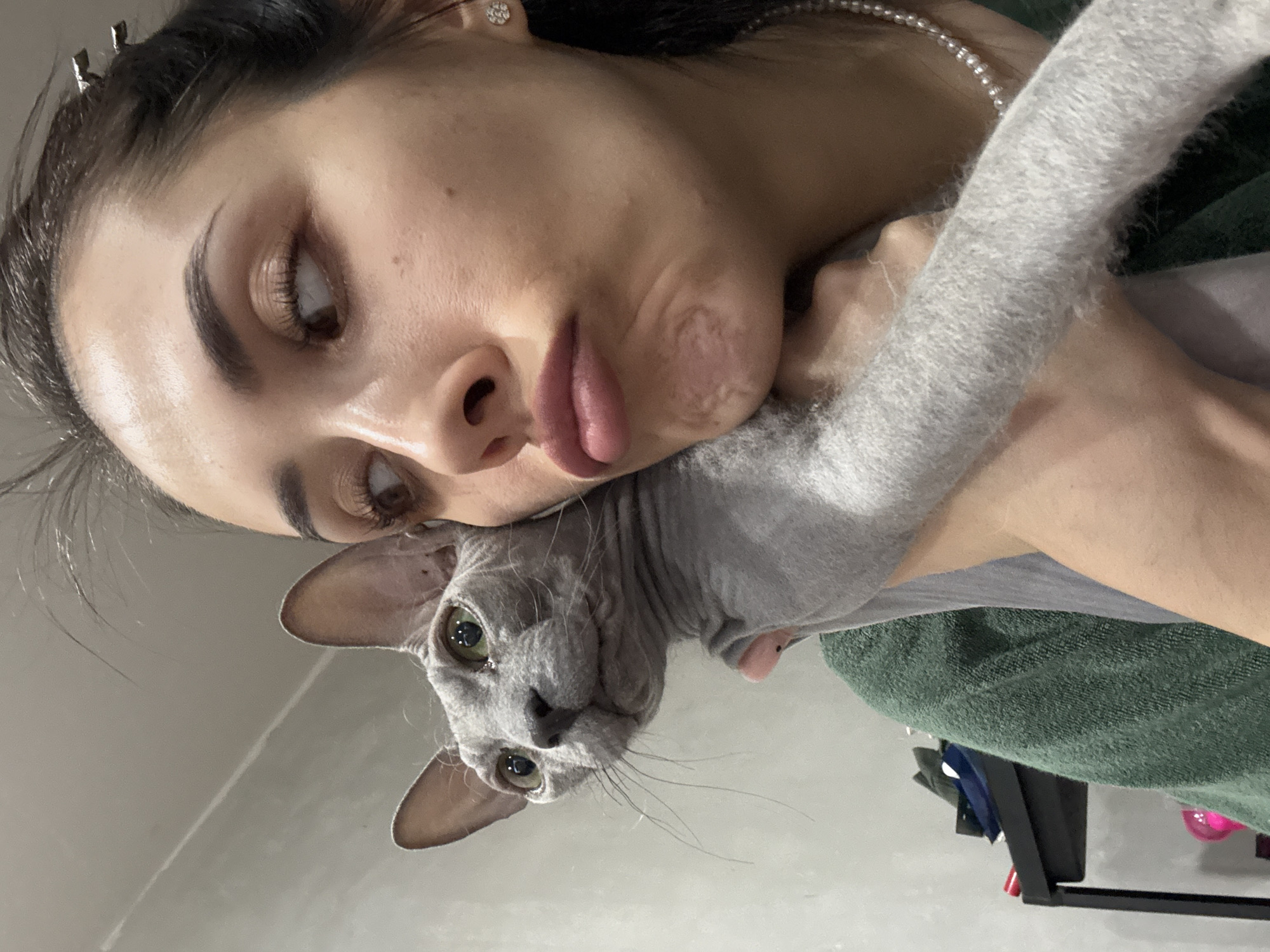 A close-up, horizontally oriented image captures a woman holding a Sphynx cat in an intimate and playful pose. The woman's face, with dark hair pulled back and secured by a silver clip, is positioned on the left side of the frame. She wears a diamond stud earring and a pearl necklace. Her eyes, with long eyelashes and well-groomed eyebrows, are directed slightly away from the viewer. Her lips are puckered into an exaggerated pout or "fish face," creating a humorous expression and slight creasing on her cheek.
Her arm extends from the left, cradling a gray Sphynx cat against her. The cat's head, featuring large, striking green eyes and prominent ears, is in the lower central part of the image, gazing directly at the viewer. Its wrinkled, hairless skin and visible whiskers are distinctive. The cat's body is draped over the woman's arm or shoulder.
The background is minimalistic, primarily a light, featureless surface that could be a wall or ceiling, with a faint linear reflection running across it. In the bottom right corner, a textured dark green fabric, possibly clothing or a blanket, is visible, along with some indistinct dark objects that appear to be part of furniture or a stand. The lighting is soft and even, indicating an indoor setting. The image conveys a sense of affection and lightheartedness between the woman and her pet. No text is visible. The context for this scene is Votkinsk, Russia.