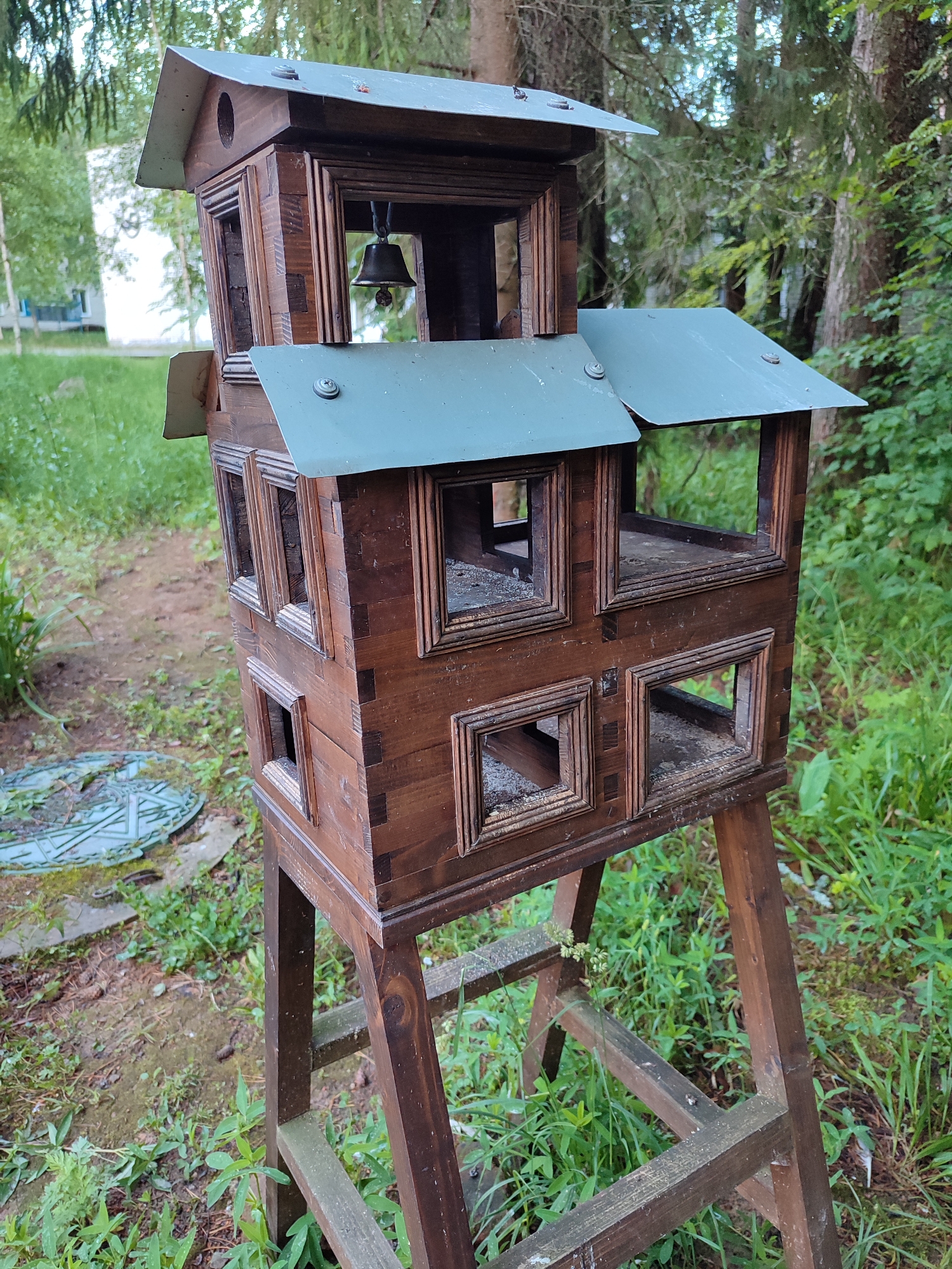 A rustic, multi-tiered wooden birdhouse with a weathered green metal roof stands on a wooden A-frame support in an outdoor, wooded environment. The birdhouse is constructed from dark brown wood, featuring multiple open window-like compartments on each level. A small brass bell hangs from the top tier. The surrounding area is overgrown with green grass and foliage, with trees visible in the background. A round, green grate is partially visible on the ground to the left of the birdhouse. The scene appears to be in a natural setting, possibly a park or backyard.