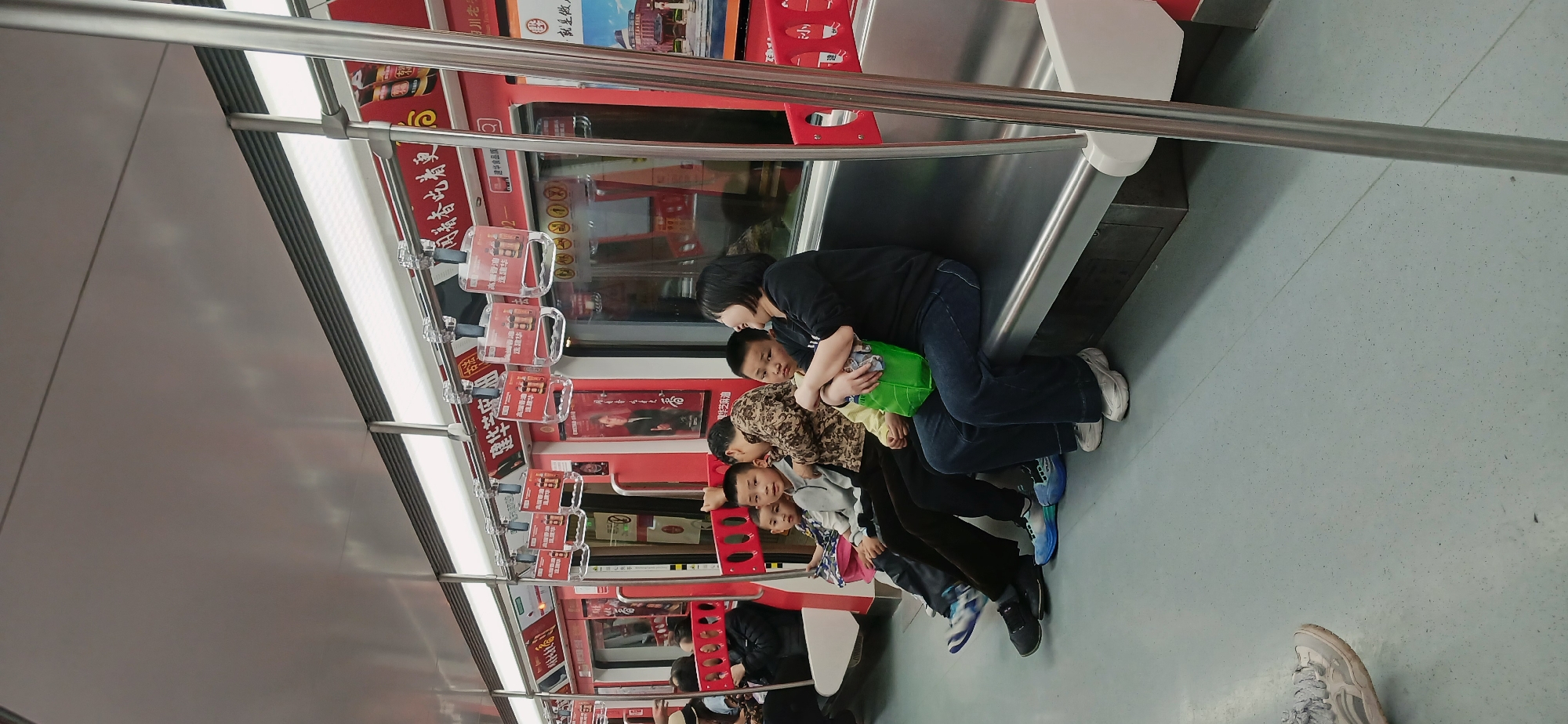 The image shows the inside of a subway car in Yuzhong District, China, with a group of people sitting along the side of the train. A woman in a black shirt and jeans is standing, bending over and seemingly interacting with a child sitting down. Three other children are also seated next to each other. The subway car has red advertisements and overhead handles. The floor of the subway car is a light gray speckled color. A metal handrail runs along the top and side of the car.