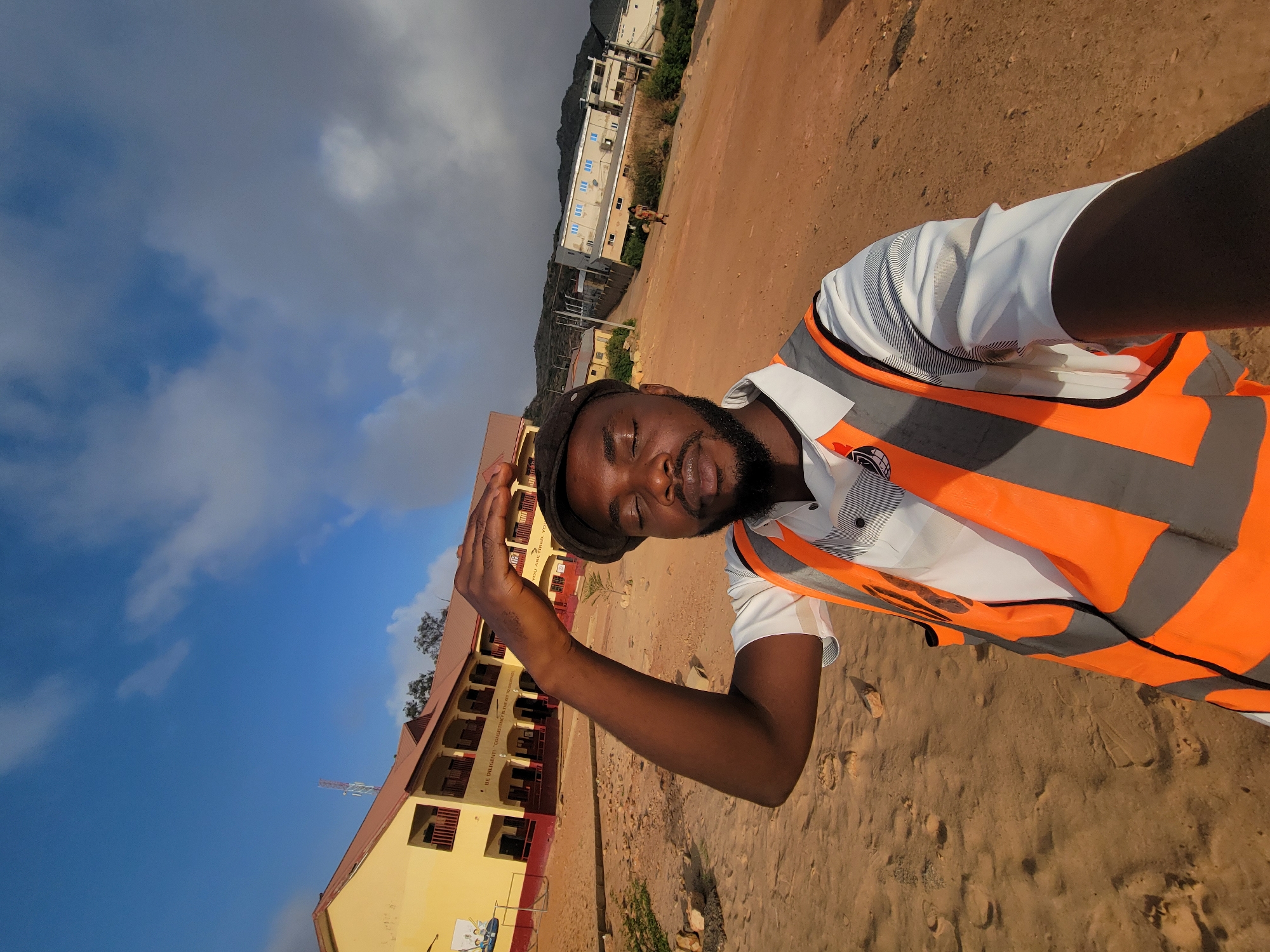 A man in a safety vest poses in front of a two-story building in Gura Top, Nigeria. He is wearing a dark cap, white shirt, and a high-visibility orange and grey safety vest. He has a beard and is touching his cap with his right hand. The building behind him is painted yellow and has a red roof. There is a metal structure extending from the roof, possibly an antenna. The ground is dirt and sand. Another building is visible in the background along with cloudy skies.