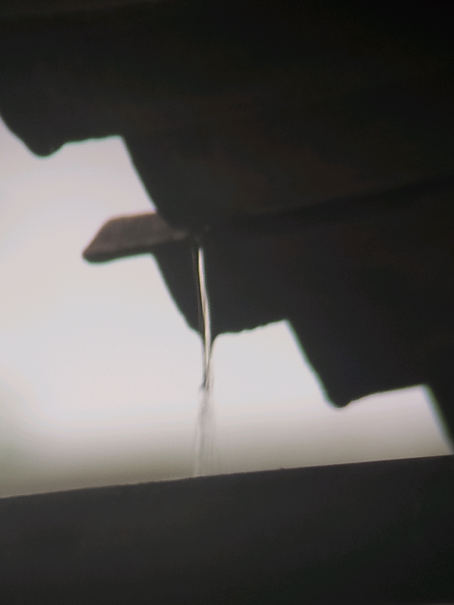The image shows a close-up view of a thin stream of liquid, likely water, dripping from a dark, angular surface against a blurred, pale background. The liquid forms a delicate, suspended droplet at its tip. The dark surface appears to be part of an overhang or architectural feature, possibly a roof or gutter. The lighting is dim, casting deep shadows. The scene is abstract and lacks any specific environmental markers that would pinpoint a location within Jember, Indonesia.