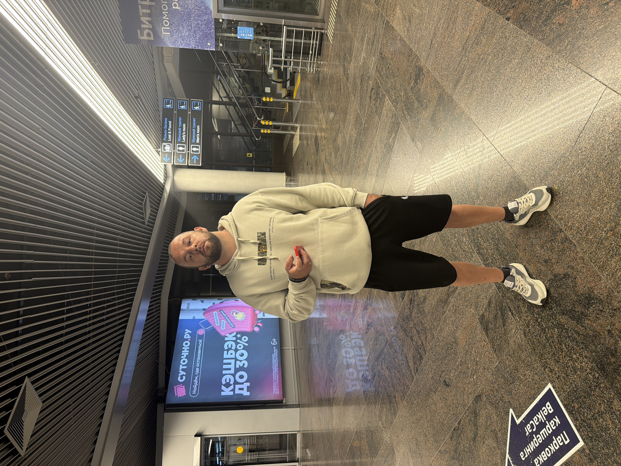 A bald white man stands in a well-lit, modern indoor space, possibly an airport or a large commercial building, with a granite tiled floor and a slatted ceiling. He is wearing a light-colored hooded sweatshirt and black shorts, along with athletic shoes and socks. He is holding a small red object in his right hand.
To his left, a large digital advertisement displays text in Russian, promoting a 30% cashback offer. To his right, a directional signpost indicates "Lost & Found", "Lady's room", and "Men's room", with corresponding arrows and pictograms. Further to the right, a blue sign with white text, partially visible, appears to be a welcome message.
On the floor, a blue and white arrow sticker with Russian text points to the left, indicating "Parking". The man's body is angled slightly towards the camera, and he looks directly at it. The lighting is bright, casting reflections on the polished floor and the man's clothing. The overall impression is one of a casual moment captured in a public transit or commercial hub, likely in Sochi, Russia, given the Russian text visible in the image.