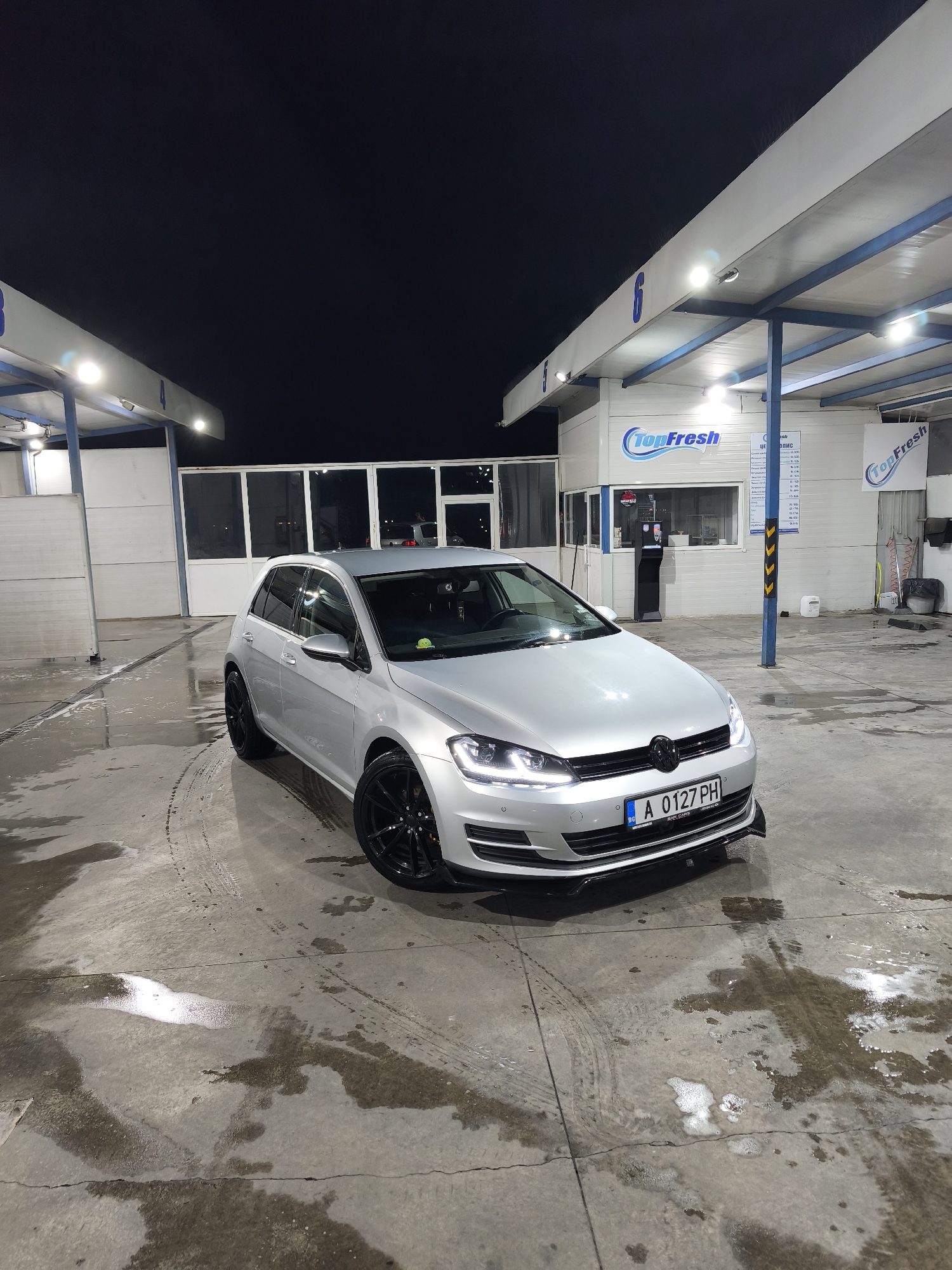 The image depicts a silver Volkswagen Golf parked at a self-service car wash in Burgas, Bulgaria. The car is positioned on a wet concrete surface with tire tracks and puddles of water and foam residue. The car wash has multiple bays under covered canopies, identified by numbers, with "TopFresh" branding. Automated payment kiosks and service equipment are visible in the background. The sky is dark, suggesting it is night time. The license plate of the car reads "A 0127 PH".