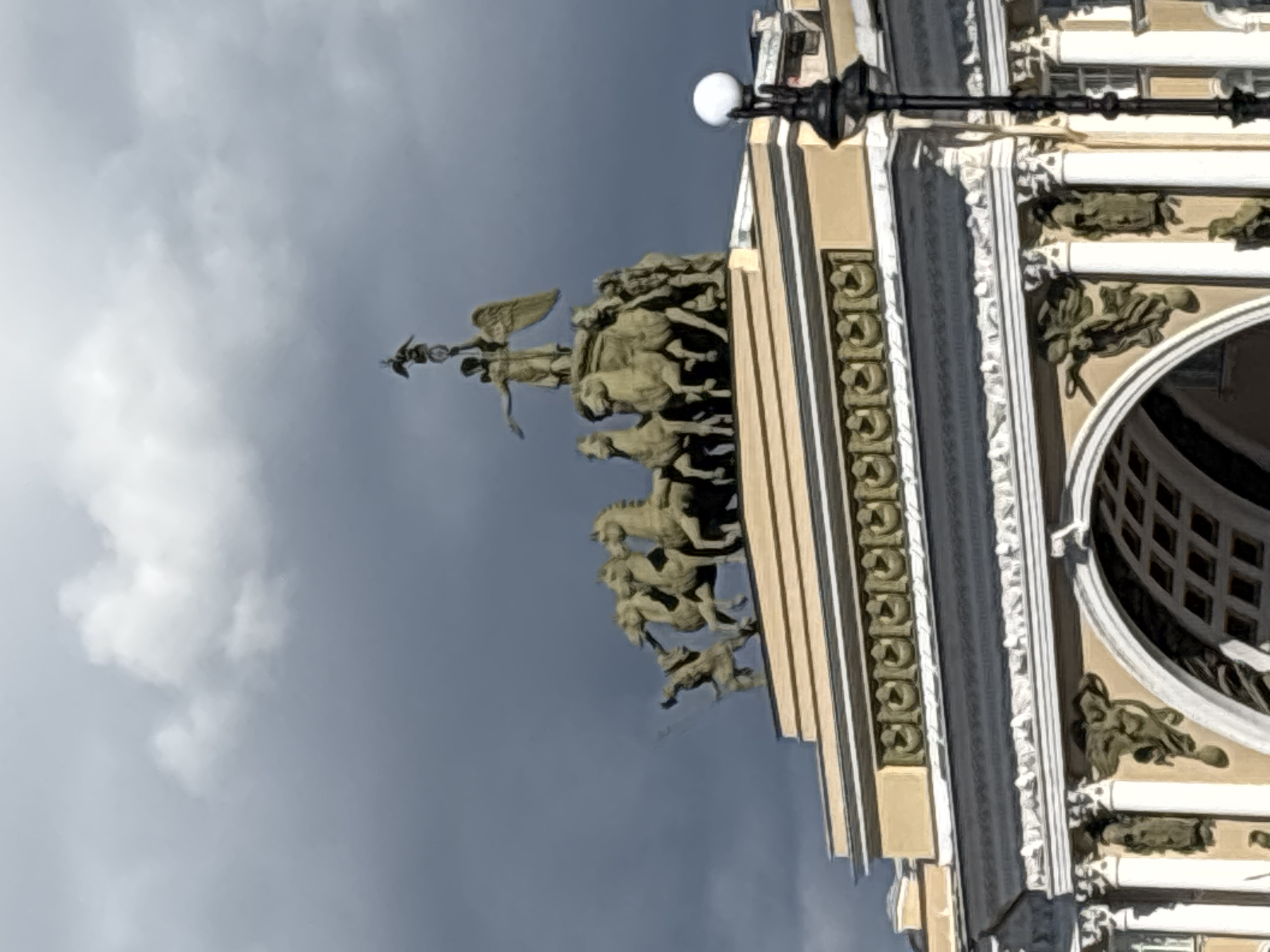The image shows the top of the Triumphal Arch of the General Staff Building in Palace Square, Saint Petersburg, Russia. The arch is crowned with a sculpture group depicting the Goddess of Victory in a chariot drawn by six horses. The sky is dramatic, with dark storm clouds contrasting with lighter patches of sky. A lamppost with a spherical light fixture is visible on the right side of the frame. The architecture of the building is ornate, with decorative carvings and columns visible beneath the sculptural group.