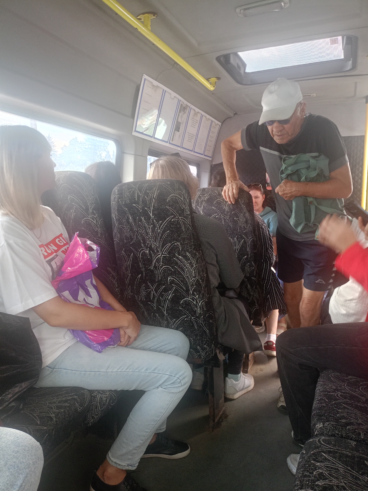 The image shows the interior of a crowded bus or shuttle van, likely in Zlatoust, Russia, given the context. Several passengers are visible, seated and standing. In the foreground, a young woman with blonde hair sits on a seat, wearing a white t-shirt and light blue jeans. She is holding a pink and purple plastic bag. Next to her, another passenger's legs are visible. In the middle ground, an older man wearing a white baseball cap, sunglasses, and a dark t-shirt is standing, holding a green bag. He appears to be interacting with someone further back. Other passengers are seated and standing in the background, with glimpses of their clothing and hair visible. The seats are upholstered with a dark fabric featuring a white leaf-like pattern. The interior of the vehicle appears functional, with overhead compartments and lighting fixtures. Natural light streams in from a window on the left, illuminating the scene. A poster or information sheet is mounted on the wall above a window. The overall impression is of a mode of public transportation during daylight hours.