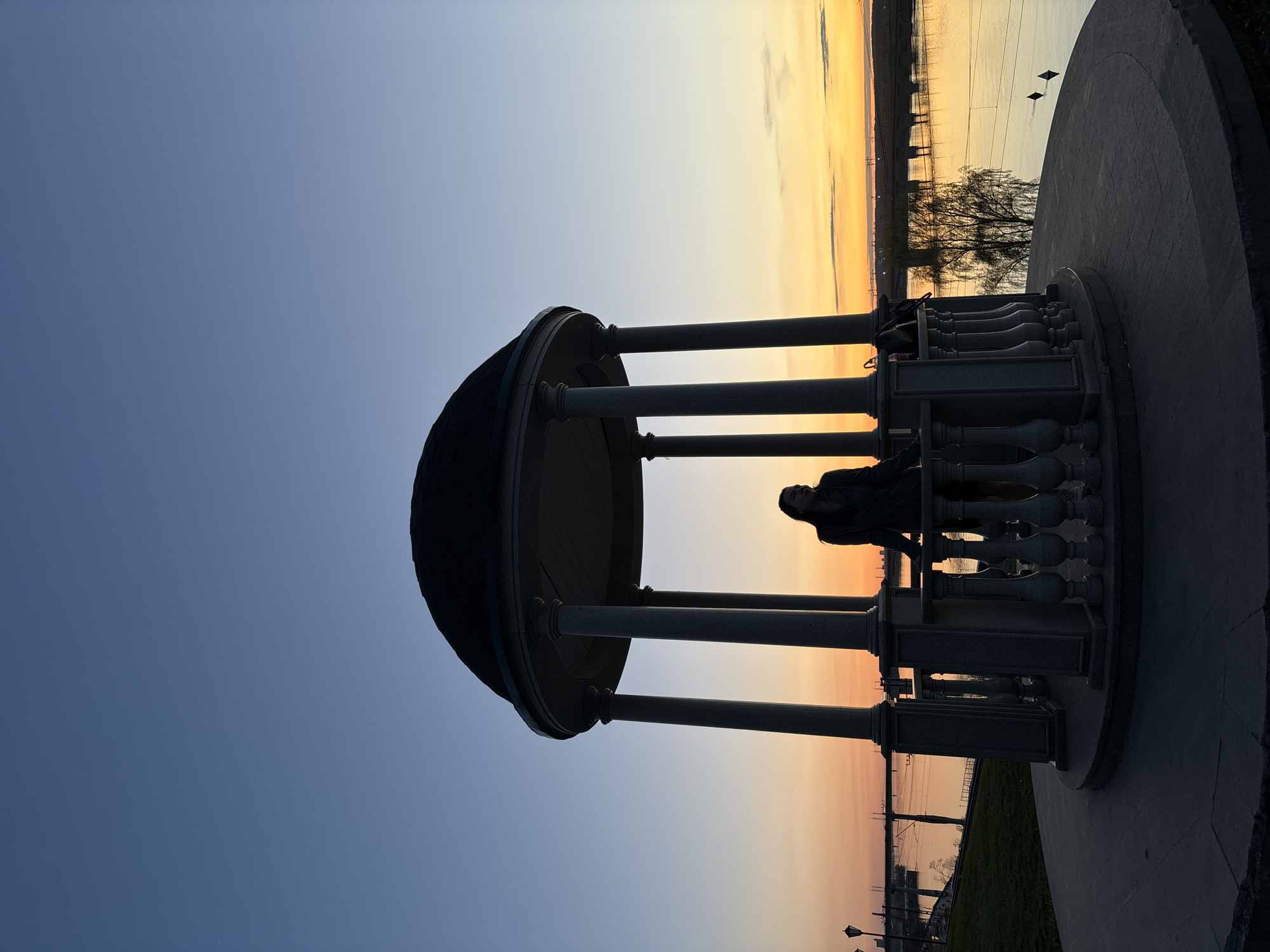 A gazebo is located on a street with a sunset in the background. A person is leaning against the inside of the gazebo. The image is rotated horizontally. The scene is in Perm, Russia.