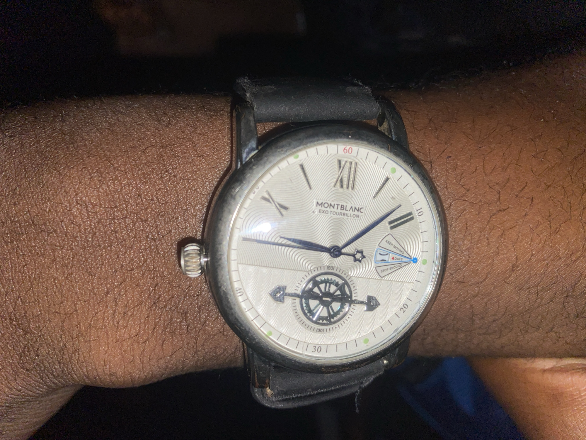 A close-up shot of a person's wrist wearing a Montblanc Exo Tourbillon watch. The watch has a silver-toned dial with Roman numerals and a sub-dial displaying a rotating mechanism with a compass-like design. The watch band is made of black leather. The person's skin is dark-skinned and has visible pores and hairs. The background is dark and out of focus, with a hint of blue and black. The location is not identifiable in the image and there is no information about the city of Wumat, Nigeria.