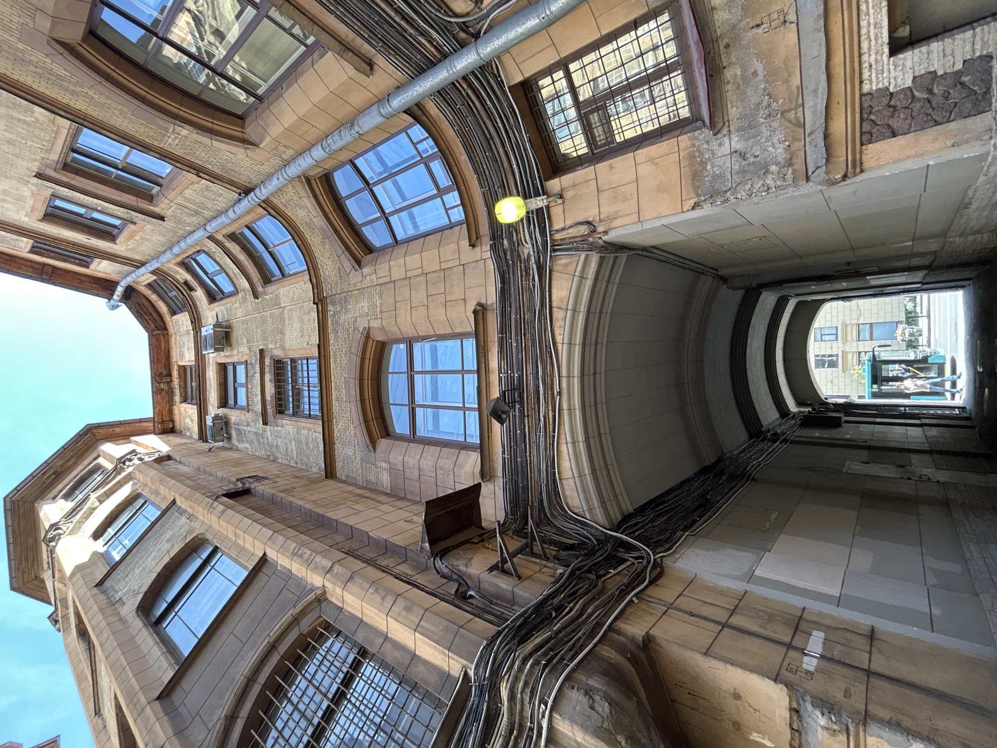 The image depicts a narrow passage between two tall, old buildings with ornate architecture, viewed from below and angled upwards towards a clear blue sky. The buildings are constructed with a combination of brick and stone, featuring numerous windows of varying sizes and styles, some with decorative grilles. A dense network of electrical wires and pipes runs along the exterior walls, creating a visually complex pattern. A single yellow streetlamp is illuminated, casting a glow on the scene.
At the far end of the passage, an open area is visible, with a bus and a few pedestrians. One person, wearing a white top and blue pants, is walking away from the camera. The lighting suggests it is daytime, with sunlight casting shadows and highlighting the textures of the buildings. The overall atmosphere is one of historical urban architecture and functional infrastructure.
