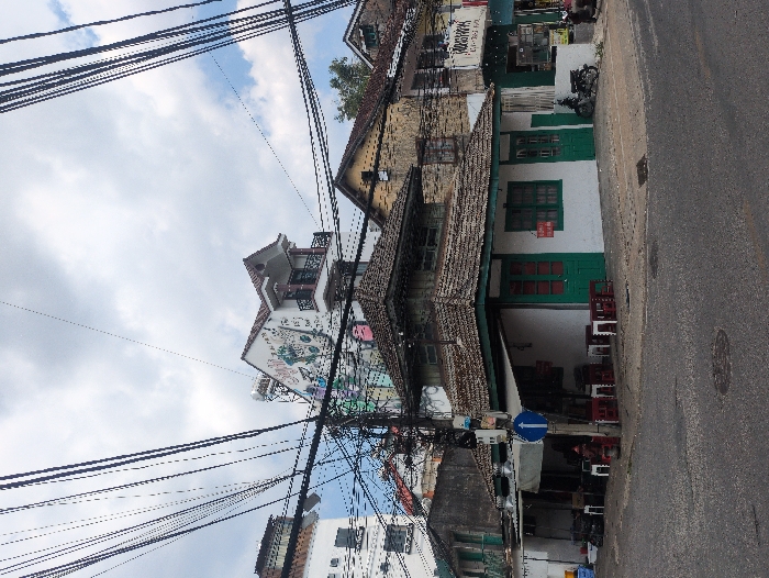 The scene depicts a street corner at 16 Lê Hồng Phong, Phường 4, Thành phố Đà Lạt, Lâm Đồng, Vietnam. A two-story building, identified as Thư Quán coffee shop, occupies the corner, featuring a white facade with dark green window frames and doors, and a multi-tiered brown tiled roof. On the ground level, several low red plastic tables with small white stools are arranged on the sidewalk under an awning.
To the left of the corner building, a weathered, unpainted two-story structure with a balcony is visible, followed by a taller white building with multiple small square windows. A utility pole stands prominently on the sidewalk, bearing a blue circular street sign with a white left-pointing arrow. A red fire hydrant is located near the pole. Various items, including blue containers, are stacked on the sidewalk next to the weathered building.
To the right, across the apparent intersection, an older, yellow-ochre building with brown windows is visible. Further right, a building with a blue facade and a partial Vietnamese sign is present. A black motorcycle is parked near the rightmost building.
The upper portion of the image is dominated by a dense, complex network of black overhead utility wires crisscrossing against a partly cloudy sky with patches of blue. The asphalt street in the foreground shows minor cracks, and a metal manhole cover is visible. No individuals are clearly identifiable or actively engaged in specific interactions; however, a faint figure is discernible in the shaded outdoor seating area to the left, and another partial figure is near the motorcycle on the far right.
