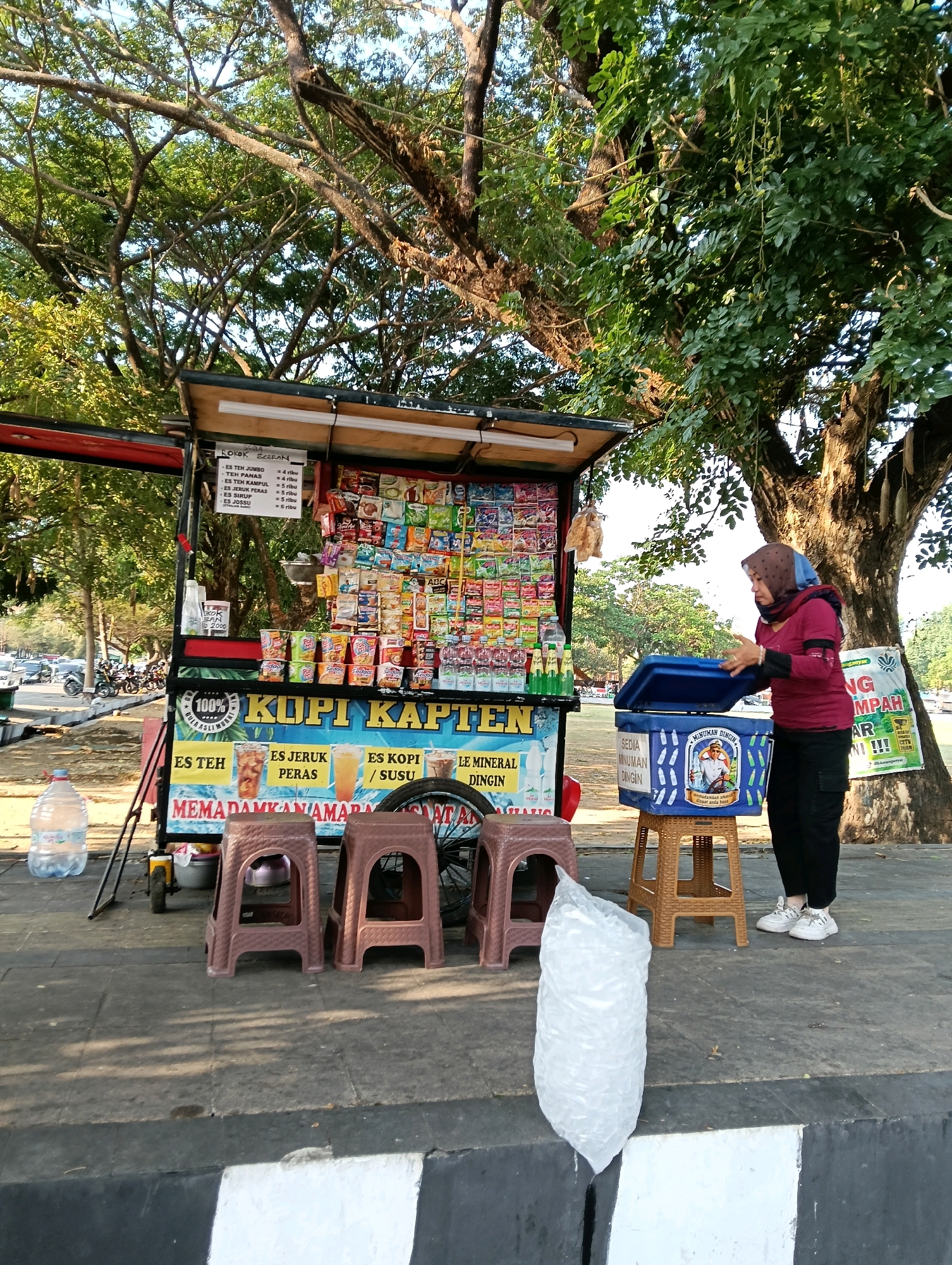 An outdoor food stall, likely operating in a park or along a pedestrian walkway in Karanganyar, Indonesia, is depicted in the image. A woman, wearing a maroon long-sleeved shirt, black pants, and white sneakers, is interacting with a blue cooler box placed on a tan stool. The cooler box has a graphic depicting a man in a captain's hat and text that reads "MINUMAN DINGIN SEDIA MINUMAN DINGIN." To the left of the cooler is a red and white food stall with various packaged snacks and drinks displayed on shelves. A sign on the stall reads "100% KOPI KAPTEN" and lists drink options such as "ES TEH," "ES JERUK PERAS," and "ES SIRUP." In front of the stall are three plastic stools and a large plastic water bottle. A bag of ice sits on the ground to the left of the stools. In the background, lush green trees with visible branches dominate the scene, with a clear sky overhead. A street with vehicles and motorcycles is visible in the distance.