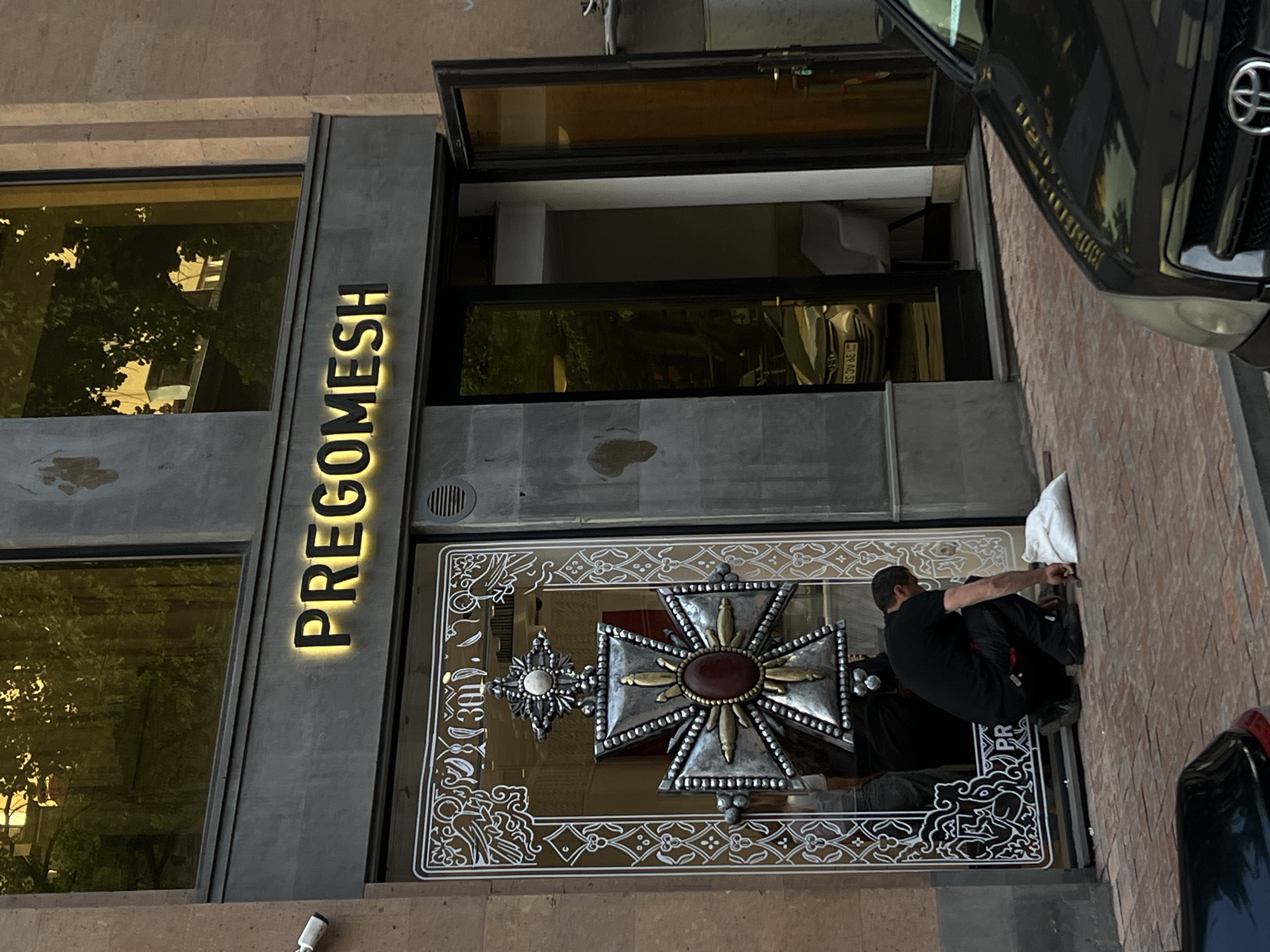 This media file presents the exterior of a building in an urban setting, identified by the illuminated vertical sign "PREGOMESH". The scene is located in Yerevan, Armenia.
The building facade features dark gray panels and large glass windows. A striking, ornate metallic decoration, resembling a large, intricate cross or emblem with silver, gold, and a reddish-brown central jewel, is prominently displayed on the glass near the entrance. The surrounding glass itself is adorned with white decorative patterns.
A person, dressed in a dark shirt and pants, is crouched on the ground near the ornate decoration, appearing to be engaged in work, possibly cleaning or adjusting something. A white cloth is visible beside them. A black car is partially visible on the right, and its reflection in the window shows part of a license plate: "92 AG 53".
Reflections in the building's windows show green foliage from trees, parts of other structures, and a minimalist interior design with a white chair. The lighting indicates it is daytime, perhaps late afternoon, as the "PREGOMESH" sign is illuminated and stands out. A security camera is visible on the lower left facade.