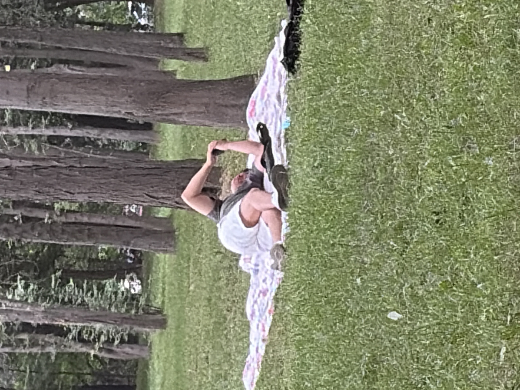 A person is lying on their back on a blanket spread on the grass, outdoors. They are wearing a dark shirt and white shorts. Their legs are bent, and one arm is extended upwards, holding their foot. The person appears to be doing some form of stretching or yoga. The environment is a grassy area with trees in the background, suggesting a park or wooded area. The lighting indicates it is daytime.