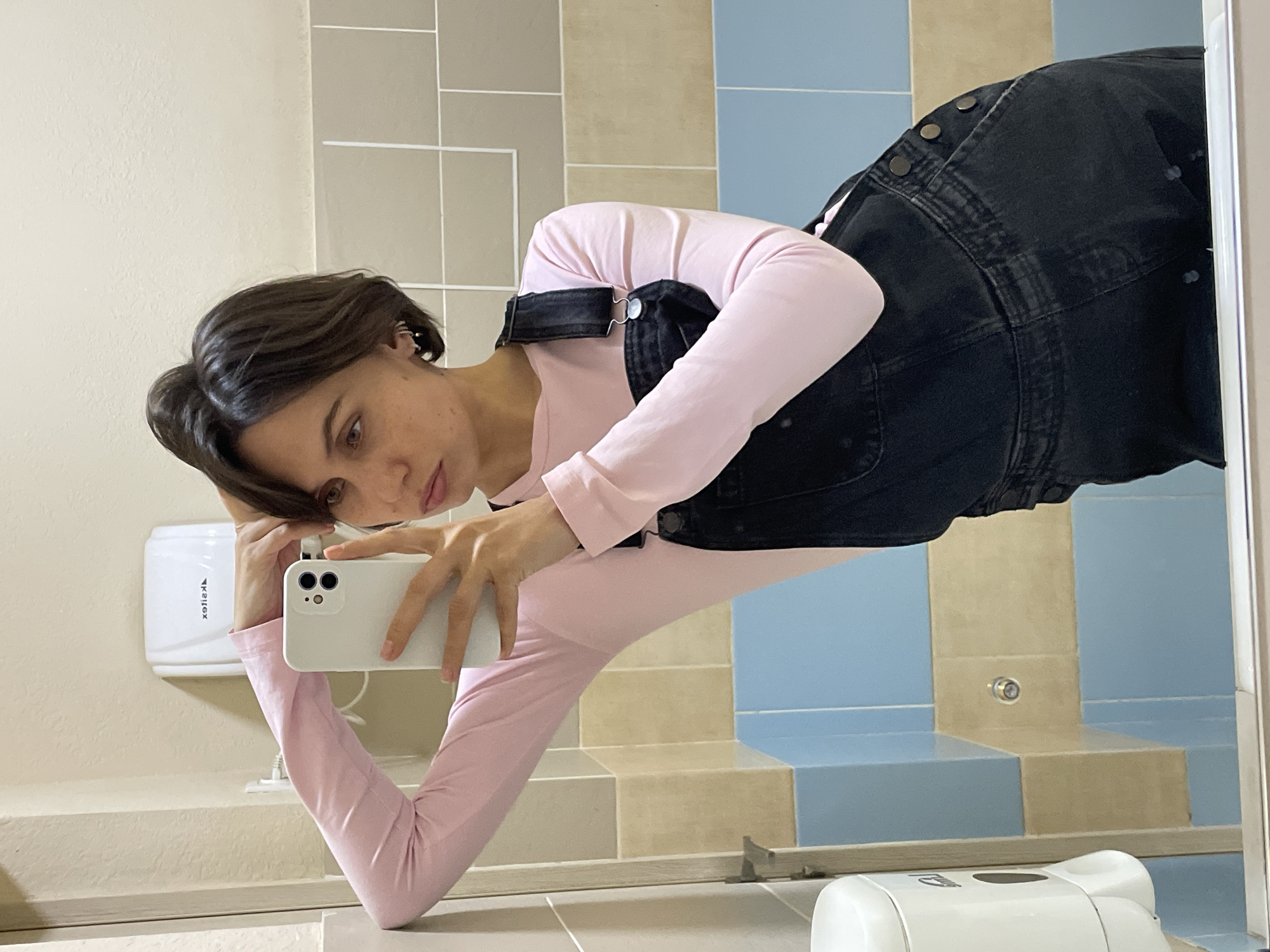 A person is captured in a mirror selfie within what appears to be a public restroom setting, in Izhevsk, Russia. The image itself is rotated 90 degrees clockwise, presenting the person and environment horizontally.
The subject is a young individual with short, dark brown hair, wearing a light pink long-sleeved top and dark denim overalls with metallic buttons and clasps. They are holding a light-colored smartphone with multiple camera lenses, looking directly into the mirror to frame the shot. A silver ear cuff is visible on their left ear. Their body is positioned at an angle, with one arm resting on a light-colored countertop or ledge.
The background consists of tiled walls with a distinct pattern of alternating light beige/cream and light blue square tiles. On the wall, above the countertop, a white hand dryer is visible, bearing the reversed text "Xeltex." Part of a white dispenser, possibly for soap or paper towels, can be seen on the countertop in the lower right of the frame. A small metallic fixture is also visible on one of the wall tiles. The overall setting suggests a utilitarian public space. The person's expression is neutral and focused on the act of taking the photograph.