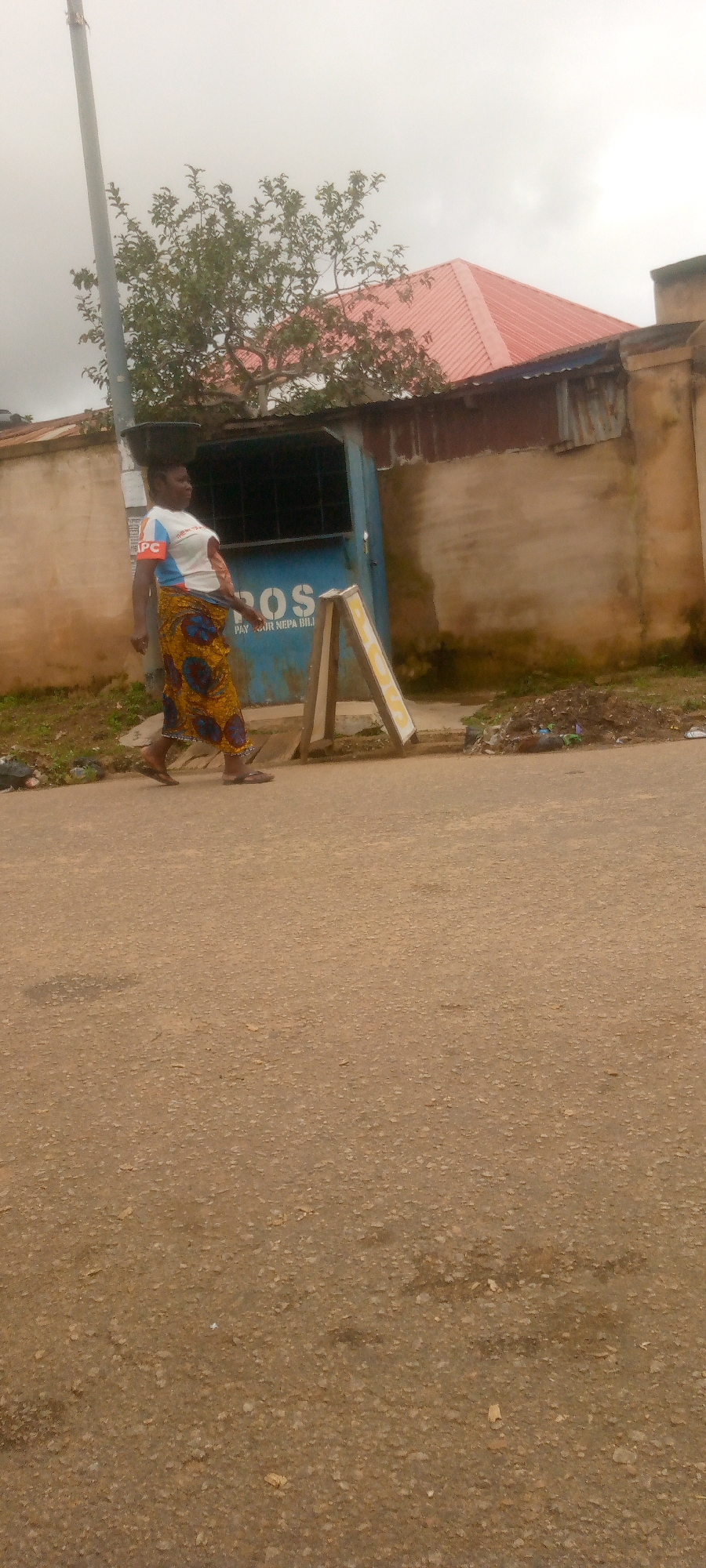A full-shot image shows a woman walking across a paved road, carrying a black bowl on her head. The woman is wearing a white shirt with blue and red accents and a vibrant yellow and blue patterned skirt. She is walking past a blue kiosk with "POS" and "PAY YOUR NEPA BILL" written on it, in front of a light brown building with a corrugated metal roof. A red-roofed building is visible behind the kiosk. There is a tree to the left of the kiosk. A wooden A-frame sign stands in front of the kiosk. The road in the foreground is paved and shows some wear. The sky is overcast.
