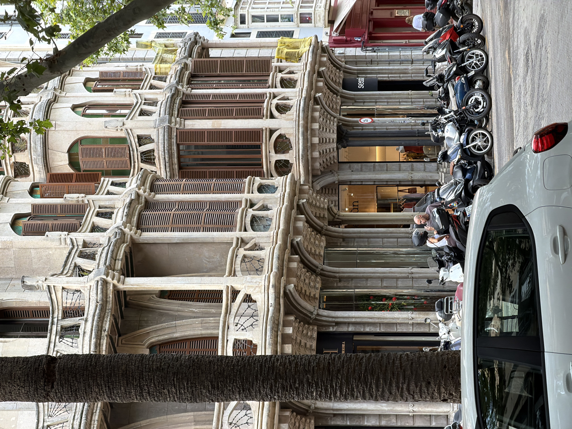 The image depicts a street scene in Palma, Spain, featuring the intricate architecture of a building with ornate balconies and shutters. In the foreground, a white car is parked on the street, reflecting the surrounding environment. Adjacent to the building, a row of motorcycles and scooters are parked along the sidewalk. Two individuals are visible near the motorcycles, one appearing to be a woman wearing a white tank top and a helmet, and the other a man in a dark shirt, both engaged in conversation. The building itself exhibits classical architectural elements, with decorative carvings and arched windows. A large palm tree trunk is prominent in the left portion of the frame, partially obscuring the building's facade. The overall atmosphere suggests a bustling urban environment on a bright day.