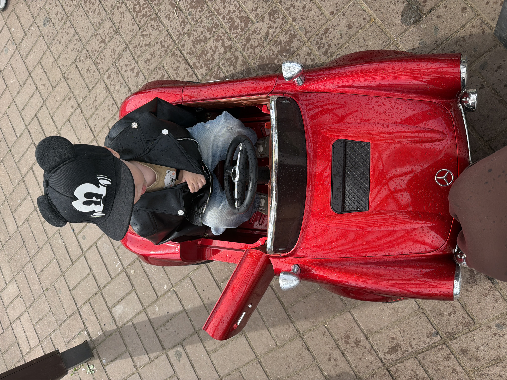 A child wearing a black baseball cap with Mickey Mouse ears and a black leather jacket sits in a shiny red toy car. The car, which appears to be a Mercedes-Benz replica, has its door open. The child is wearing blue jeans and a t-shirt with a bear graphic. The car is parked on a brick-paved area, possibly a sidewalk or a park path, in Chaikovsky, Russia. The pavement is wet, indicating recent rain. A dark-skinned hand is visible in the lower right corner of the frame, reaching towards the car.