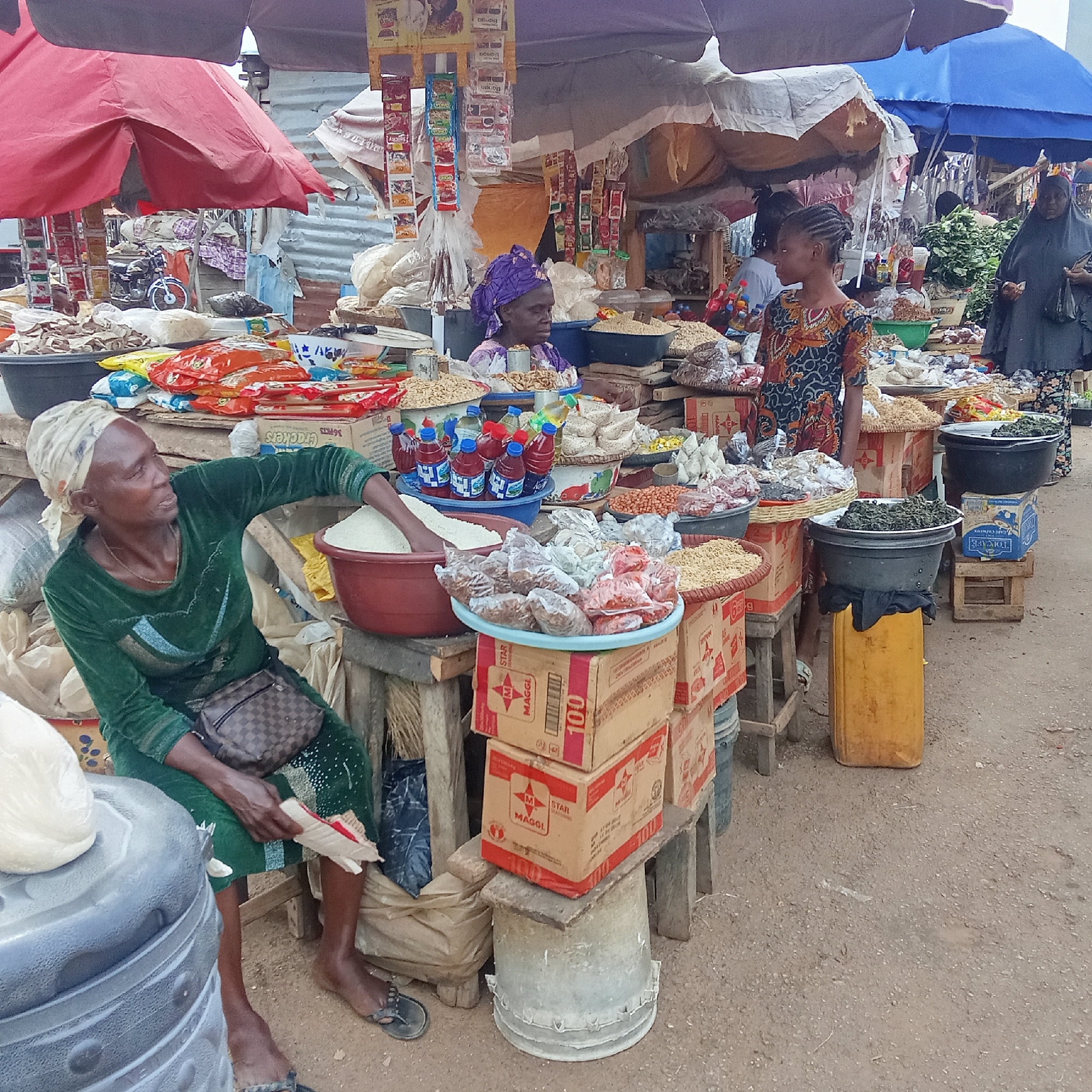 This image captures a vibrant, open-air market scene in Akwanga, Nigeria, bustling with activity under a cloudy sky or possibly shaded by large umbrellas. The ground is a mix of dirt and gravel.
Several people are visible, mostly women engaged in trading. In the foreground on the left, an older woman with dark skin and a light headscarf sits on a low stool, wearing a green long-sleeved dress and black sandals. A brown patterned bag is slung across her body. She holds small, wrapped items, possibly snacks, while beside her is a large red bowl filled with white grains, likely rice.
To her right, behind a stall laden with goods, another woman in a purple headwrap and patterned top looks towards the right. Further back, a young girl with braided hair, wearing a colorful patterned dress, stands looking left. In the background on the far right, a woman in a black abaya or long dress is partially visible, appearing to be another market-goer or vendor.
The stalls are overflowing with a variety of food items. Numerous bowls and plastic basins hold different grains, nuts (like groundnuts/peanuts), and possibly dried goods. Small plastic bottles, some containing red liquid and others clear oil, are neatly arranged. Packaged snacks and seasonings are hung from stall structures, with visible text including "Banga," "Spicity," and "TOPCAFE." Cardboard boxes, some branded "MAGGI" and "STAR MAGGI 100," are used as stands for displaying goods. Black bowls filled with leafy green vegetables are also prominent. A motorcycle is partially visible under one of the market umbrellas in the background. The scene depicts a busy marketplace where daily transactions of essential food items occur.