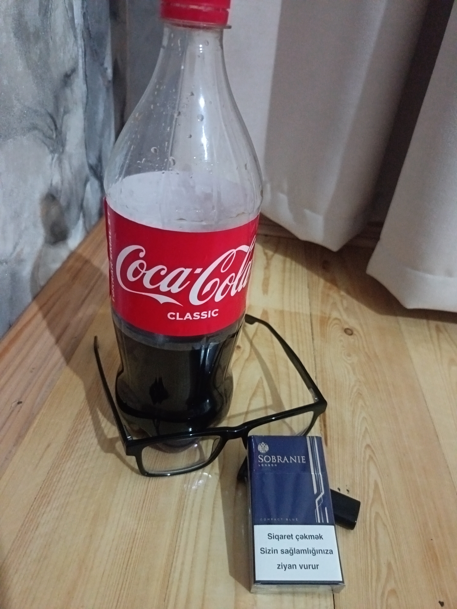 The image displays a table with various items, including a bottle of Coca-Cola Classic, a pair of black-rimmed glasses, and a pack of Sobranie cigarettes with a lighter. The Coca-Cola bottle is partially filled with a dark liquid and has condensation on its surface. The glasses are placed in front of the bottle, and the cigarette pack is positioned to the right of the glasses. The background consists of a textured wall and curtains. The overall scene appears to be indoors, on a wooden surface. There are no people visible in the image. The provided crops offer close-up views of the Coca-Cola bottle label, the cigarette pack, and the glasses. There is no information in the image that could place the scene in Beyləqan, Azerbaijan.