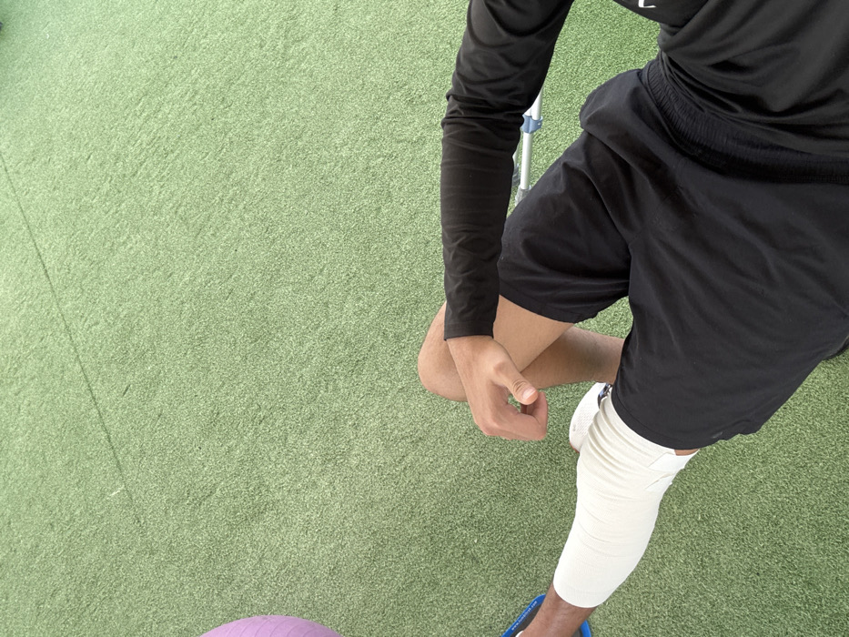 An overhead shot displays a person on a green surface, likely artificial turf. The individual wears black long sleeves, black shorts, and white athletic shoes. Their right leg is heavily wrapped with white tape or bandages from the ankle to the upper thigh. A purple exercise ball is partially visible in the bottom left corner. The person's left hand rests on their right thigh.
