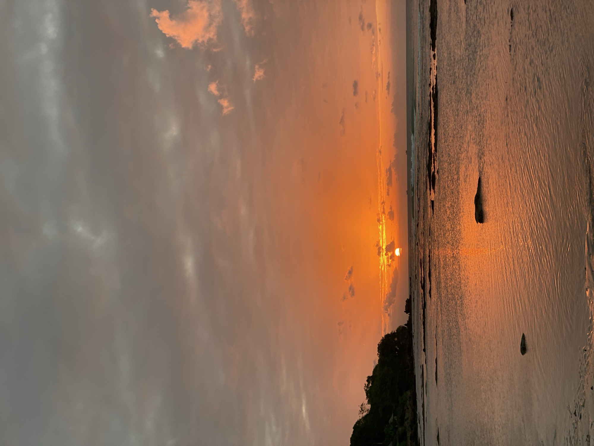 This is a vertical, candid shot of a beach during sunset. The sky is filled with dramatic, fiery orange and pink clouds, with the sun peeking through the horizon, casting a warm glow on the scene. The ocean reflects the vibrant colors of the sky, creating a shimmering surface. In the foreground, the wet sand on the beach displays ripples and textures from the receding tide, with a few dark rocks scattered across it. To the left, a lush, green hillside is visible, partially silhouetted against the bright sky. The overall mood is peaceful and awe-inspiring, capturing the serene beauty of a tropical sunset. The image appears to be taken in Kuta Selatan, Indonesia, a location known for its stunning coastal landscapes and vibrant sunsets. The time of day is clearly sunset, given the sun's position and the intense colors in the sky. The weather appears clear, with scattered clouds contributing to the picturesque scene. There is no visible text in the image.