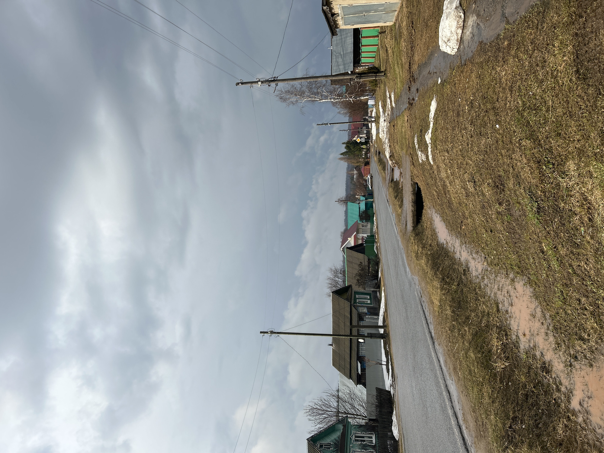 The image shows a street in Уральское, Russia. The street is paved and runs straight through a residential area. Houses line the road, most appearing to be one or two stories with traditional designs. The houses vary in color, with some painted green, white, or brown. Overhead power lines supported by wooden poles run along the street. The sky is overcast with gray clouds. Patches of snow remain on the grassy areas alongside the road, indicating it is likely early spring or late winter. The grass is brown and there are some muddy areas, suggesting recent snowmelt or rain. There is no visible human activity in the scene.