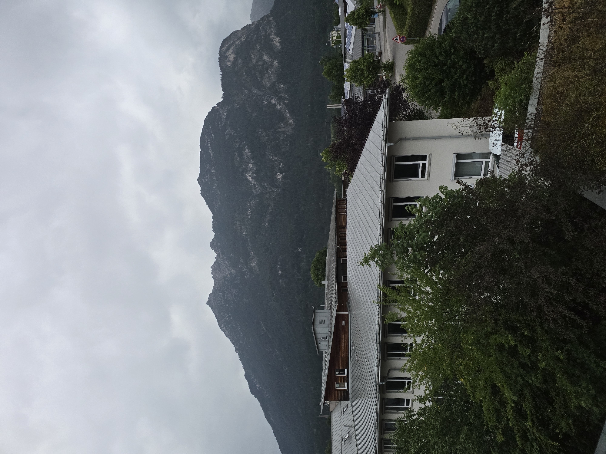 The image shows a cloudy day in Garmisch-Partenkirchen, Germany, with a large mountain range dominating the background. In the foreground, there are several buildings with grey, metal roofs and white walls. Some of these buildings appear to be offices or institutional buildings, with rows of windows visible. Trees and lush greenery surround the buildings, with some trees having dark red foliage. A road is visible in the mid-ground, with a few cars, traffic signs, and a light pole. The overall scene is serene and suggests a somewhat overcast, possibly rainy, day in a mountainous region with urban development.