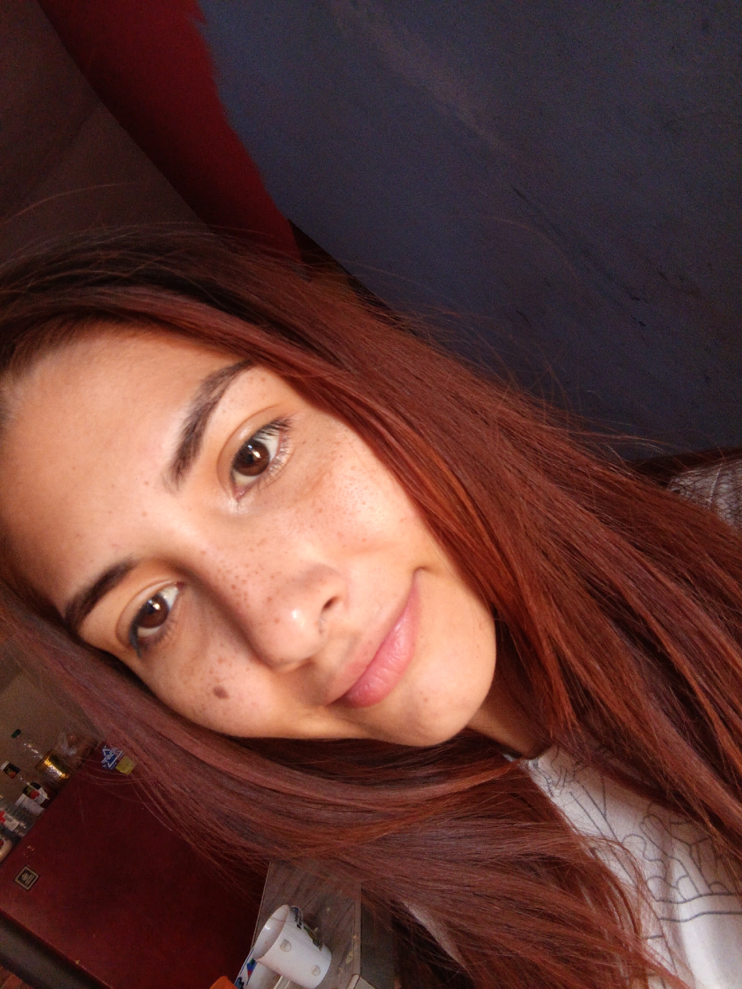 The image depicts a close-up selfie of a young woman with auburn hair. She has fair skin with freckles and a mole on her left cheek. The background appears to be an indoor setting with dark walls. In the lower left corner, there is a shelf with various items including bottles and a container. The location, Juan Griego, Venezuela, cannot be verified based on the visual information provided in the image.