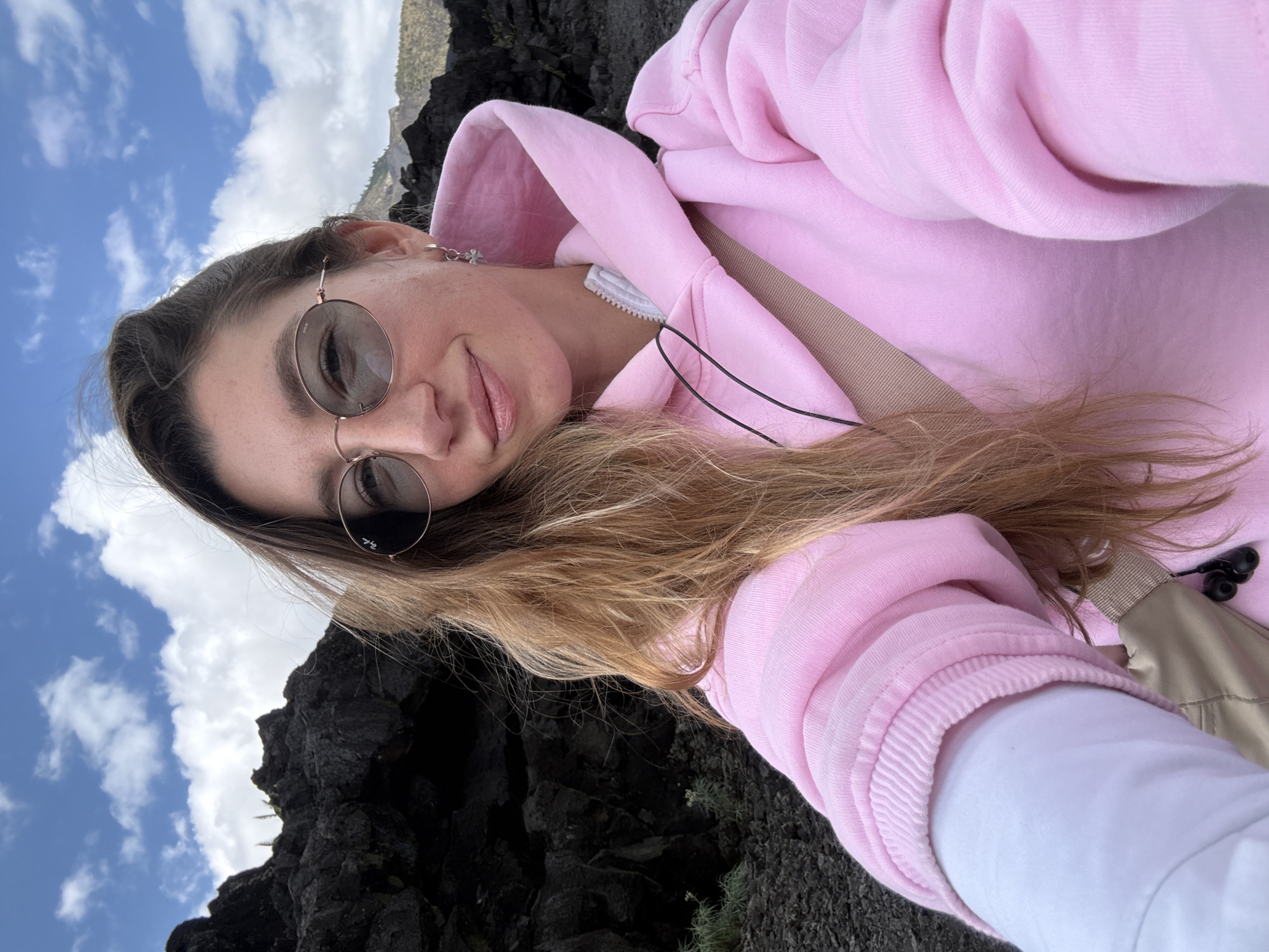 A young woman with long, wavy blonde hair, wearing round sunglasses and a pink hoodie, takes a selfie in an outdoor environment. She is smiling and looking at the camera. Behind her, dark, rocky terrain is visible, along with a cloudy blue sky. A beige strap is visible across her chest, and a white layer of clothing is visible beneath the hoodie. The scene appears to be a natural, possibly volcanic landscape. The location is identified as Kintamani, Indonesia.